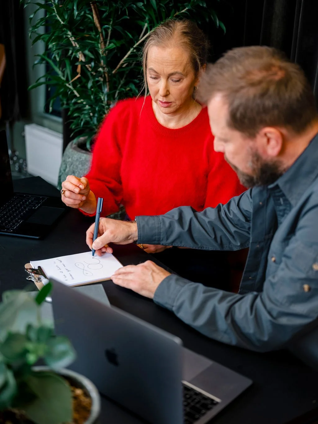 En kvinne i rød genser snakker med en mann som skriver på en notatblokk, foran en datamaskin, i et kontor med grønn plante i bakgrunnen.