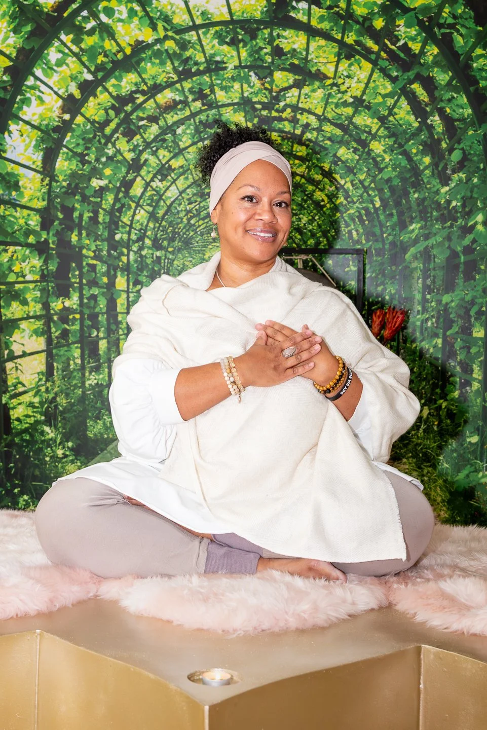 A woman sitting cross-legged on a fluffy pink mat with her hands in a prayer position, smiling in front of a green leafy tunnel backdrop.