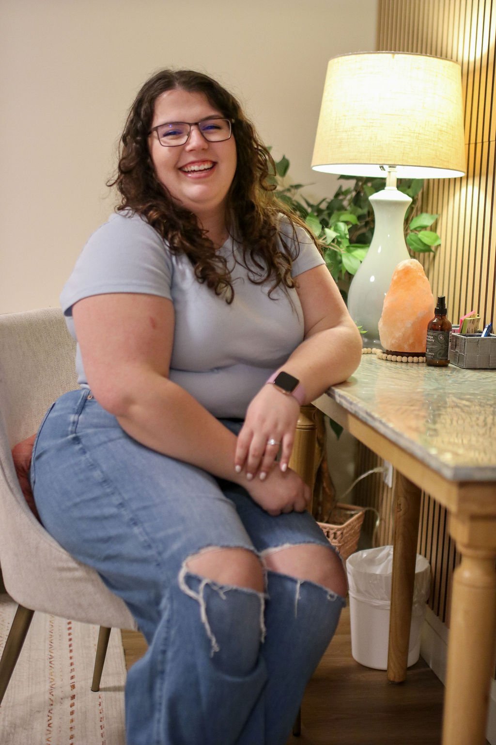 A young woman with curly brown hair and a white t-shirt sitting on a beige couch indoors, smiling at the camera.