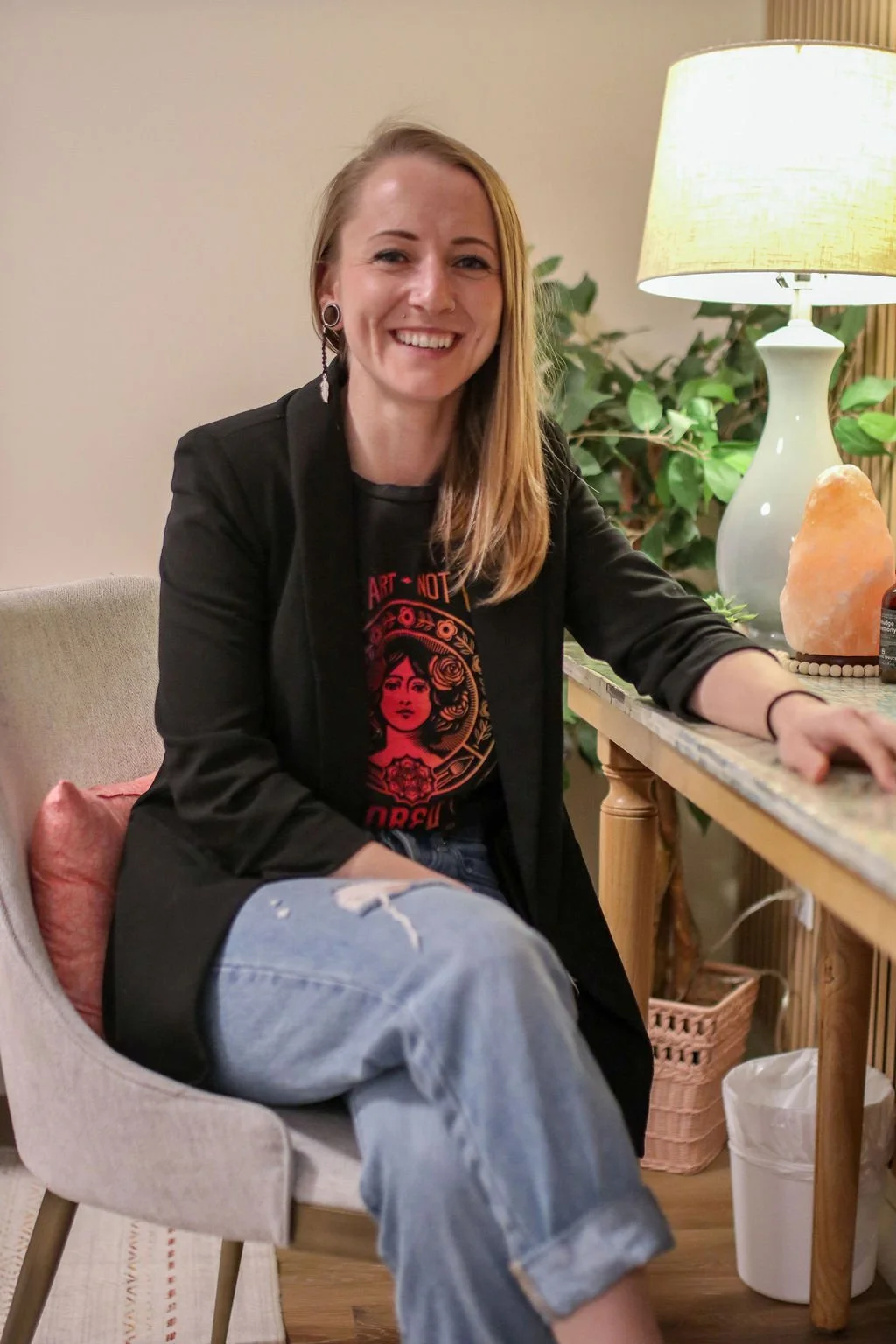 Smiling woman with shoulder-length wavy blonde hair, wearing a black shirt and knitted cardigan, sitting on a beige couch in a cozy room with a green wall, lamp, clock, and abstract art on the wall.
