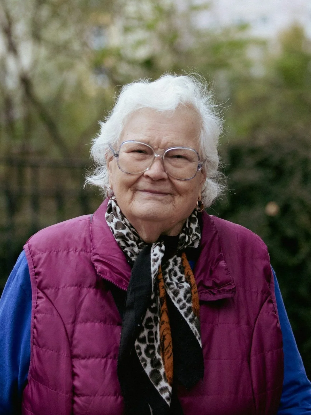 An elderly woman with white hair and glasses, smiling outdoors with blurred trees in the background, wearing a purple vest and a patterned scarf.