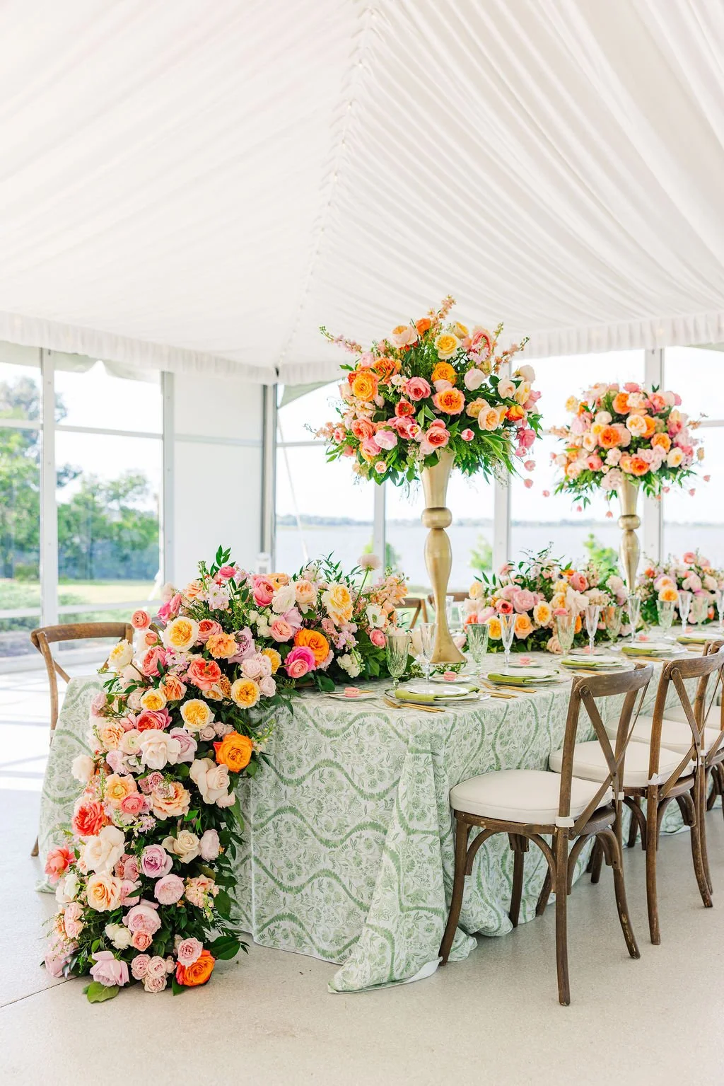 An elegantly decorated wedding reception table with large floral arrangements in tall vases, surrounded by chairs and set with plates, glasses, and cutlery inside a white tent with large windows and a view of the outdoors.