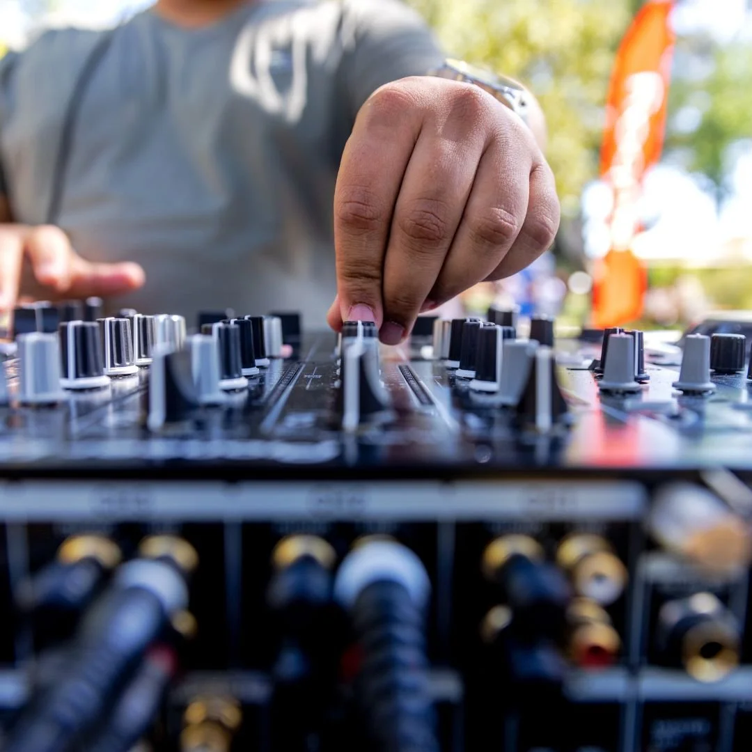 Close-up of a DJ's hand adjusting knobs on a mixing console outdoors with trees and a blurred orange flag in the background.