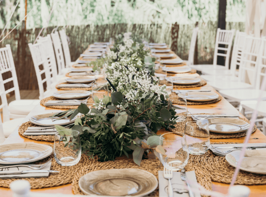Long wooden table set for a celebration with white chairs, green foliage and flowers as centerpiece, plates, glasses, utensils, and woven placemats in an outdoor setting.