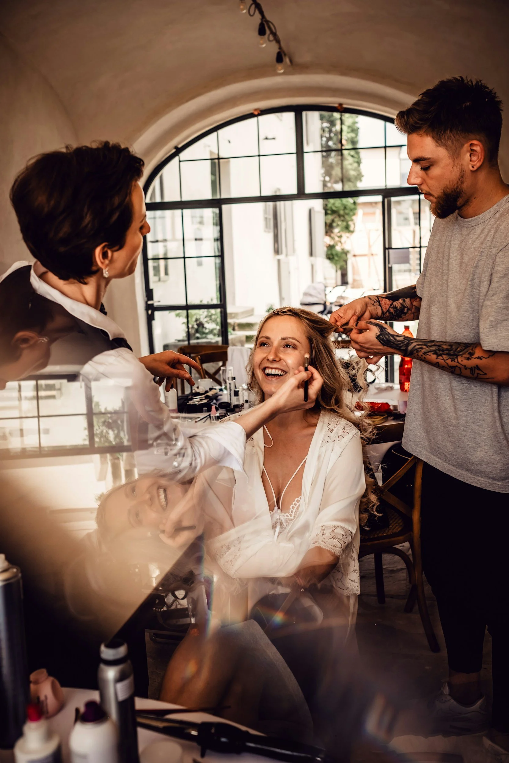 A woman getting her makeup done by a makeup artist in a well-lit room, with another stylist working on her hair, during a daytime event or photoshoot.