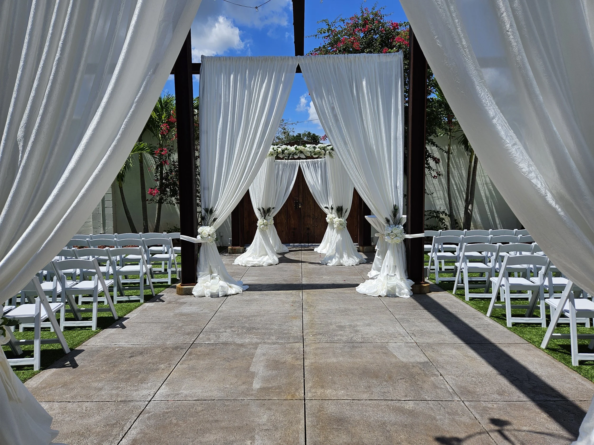 Outdoor wedding ceremony setup with white draped fabric, chairs, and floral arrangements, under a blue sky with clouds.