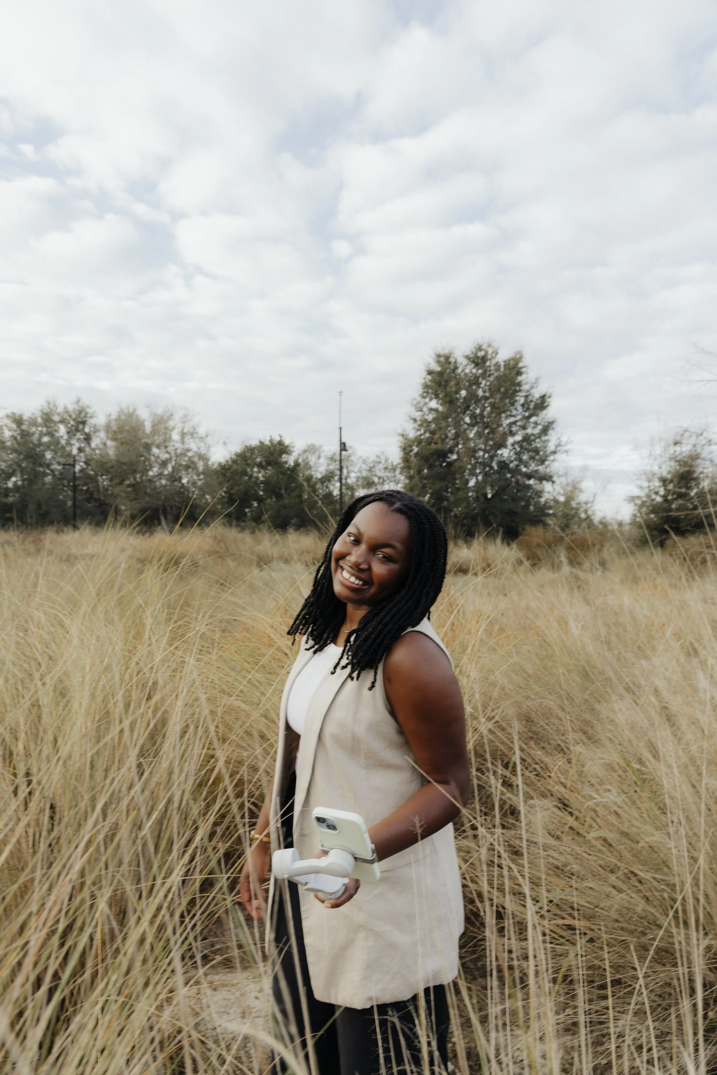 Smiling woman with dreadlocks in a beige vest holding a smartphone with a gimbal, standing in tall dry grass with trees and cloudy sky in the background.