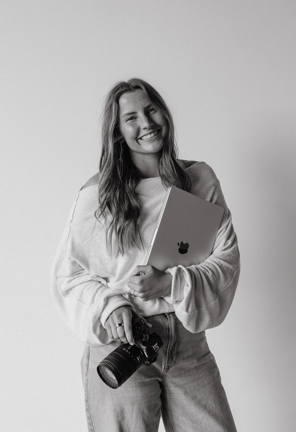 A smiling woman with long hair wearing a loose sweater and jeans, holding a camera in one hand and a laptop in the other, standing against a plain background.
