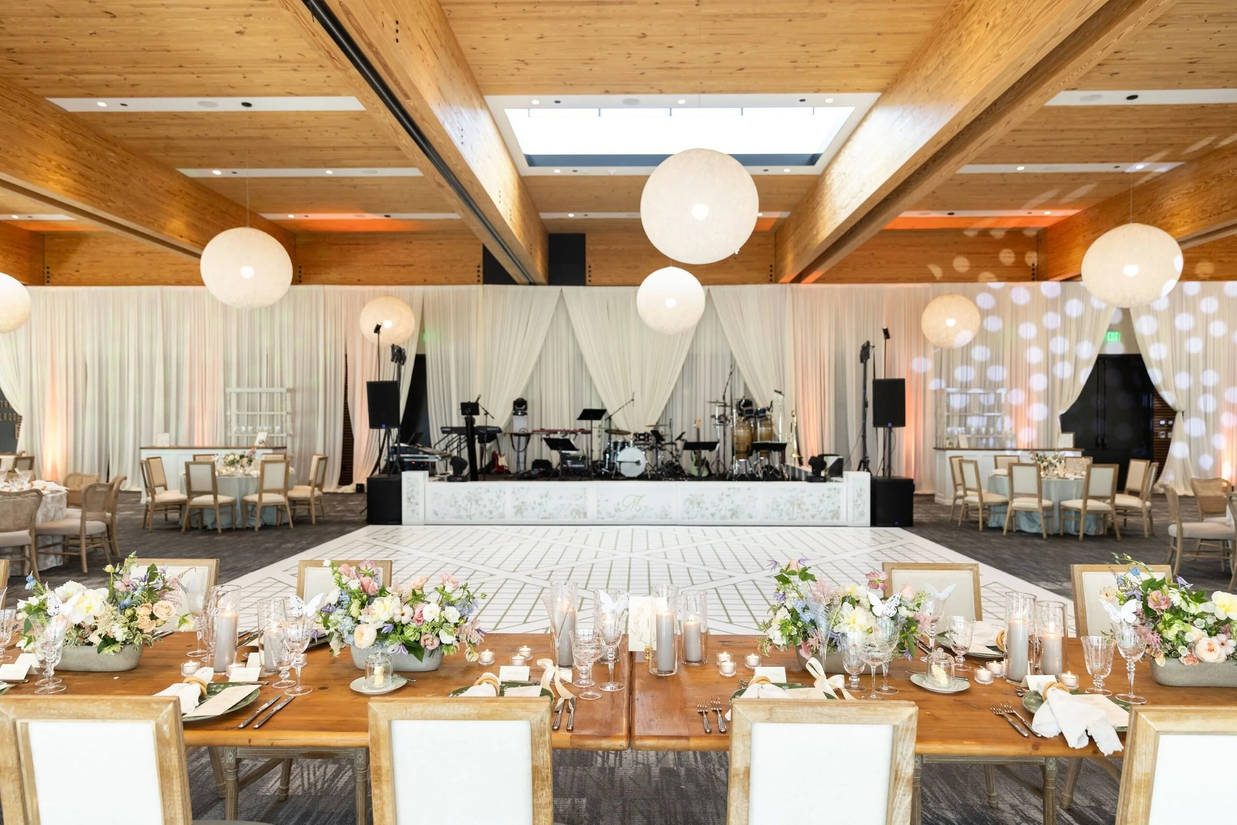 Elegant indoor event space with a stage featuring musical instruments, round tables with chairs, and a front table decorated with floral arrangements and candles, illuminated by large hanging paper lanterns.