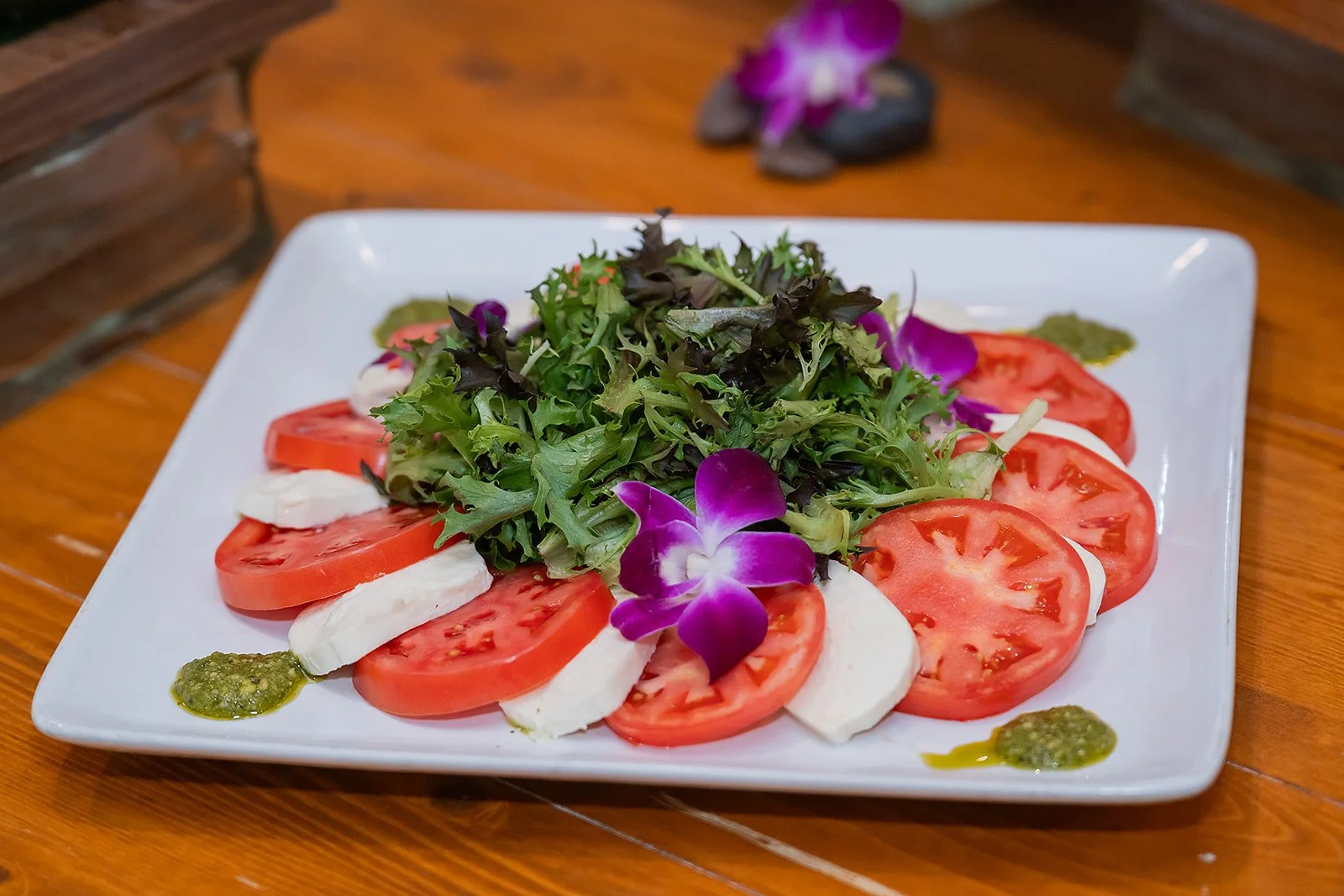 Salad with sliced tomatoes, mozzarella cheese, mixed greens, purple flowers, and green dressing or sauce on a white square plate.