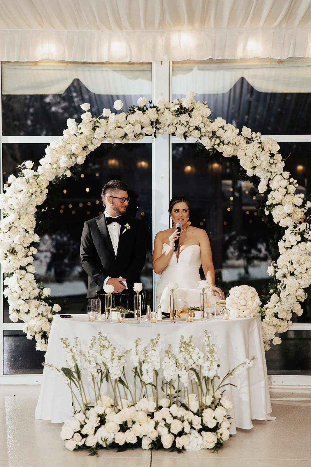 Bride and groom at wedding reception, standing behind a decorated table, with a floral heart-shaped arch behind them. The bride holds a microphone, dressed in a strapless white wedding gown, and the groom in a black tuxedo with a bow tie.