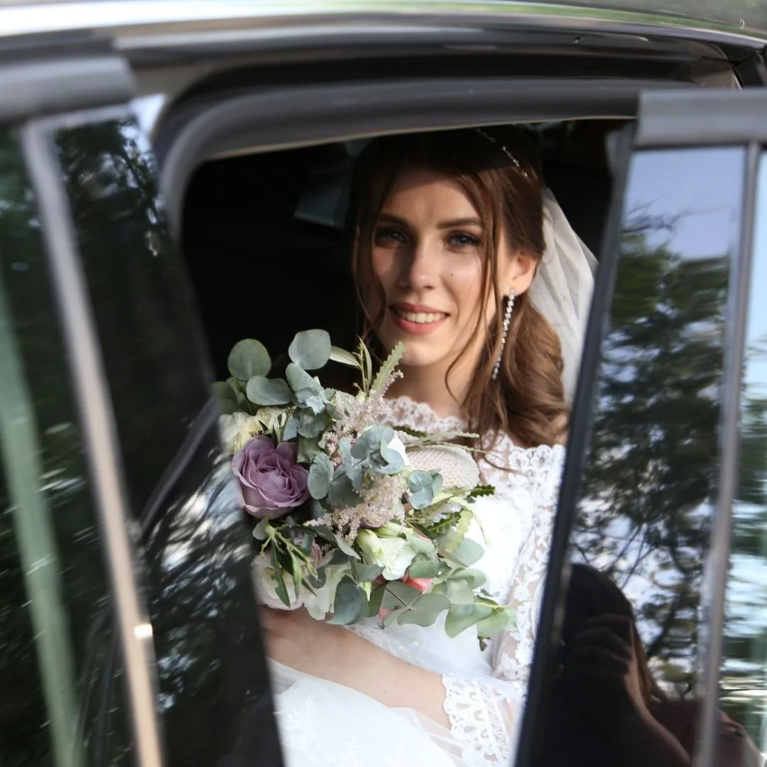 A bride sits inside a car holding a bouquet of flowers, smiling and looking out of the window.