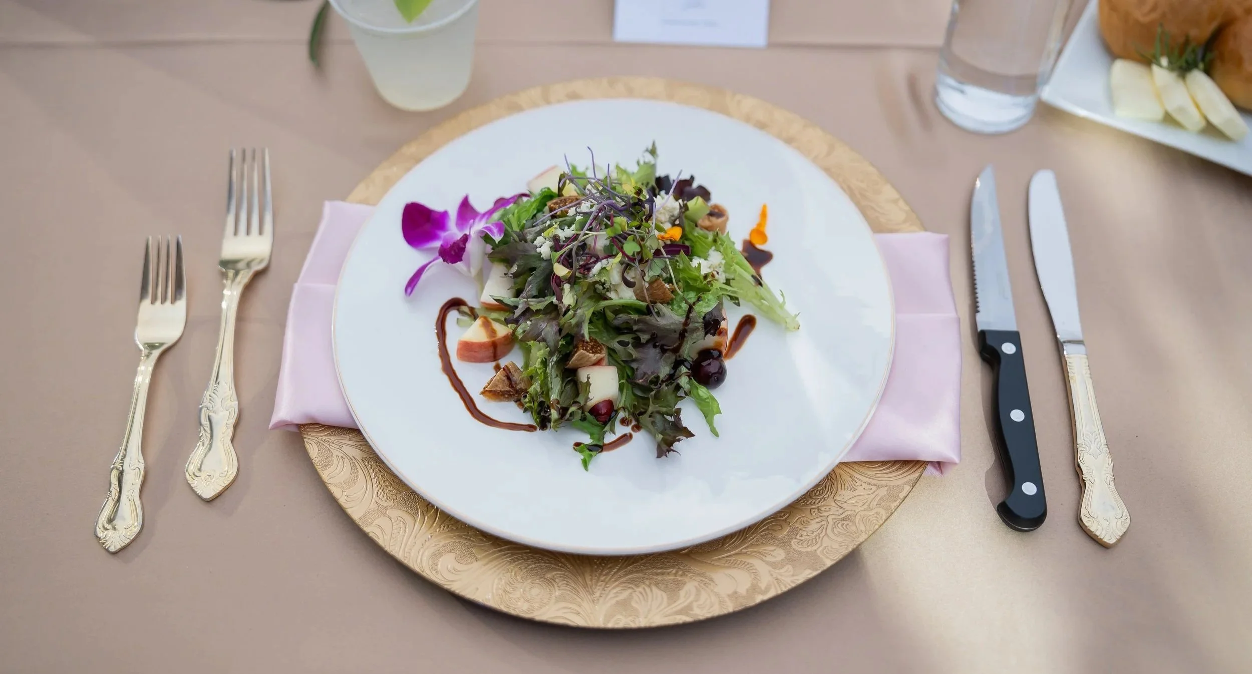 A white plate with a colorful salad, garnished with purple flowers and microgreens, on a gold charger plate with a pink napkin underneath. The table setting includes three forks on the left, a large knife and spoon on the right, and a glass of water and a small drink nearby.