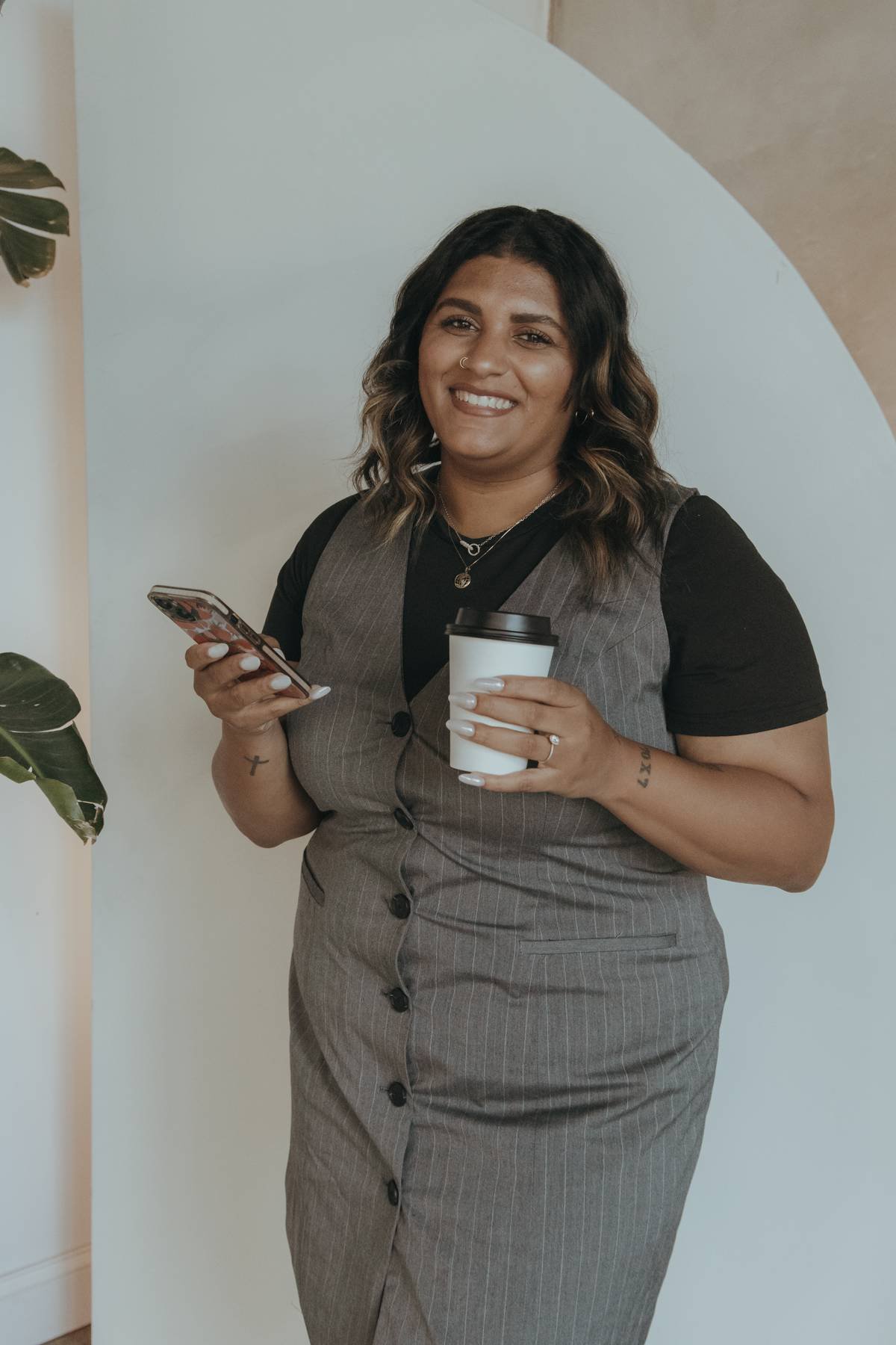 A woman with wavy dark hair holding a smartphone in one hand and a coffee cup in the other, standing indoors with a large green plant nearby and a beige wall with an archway in the background.