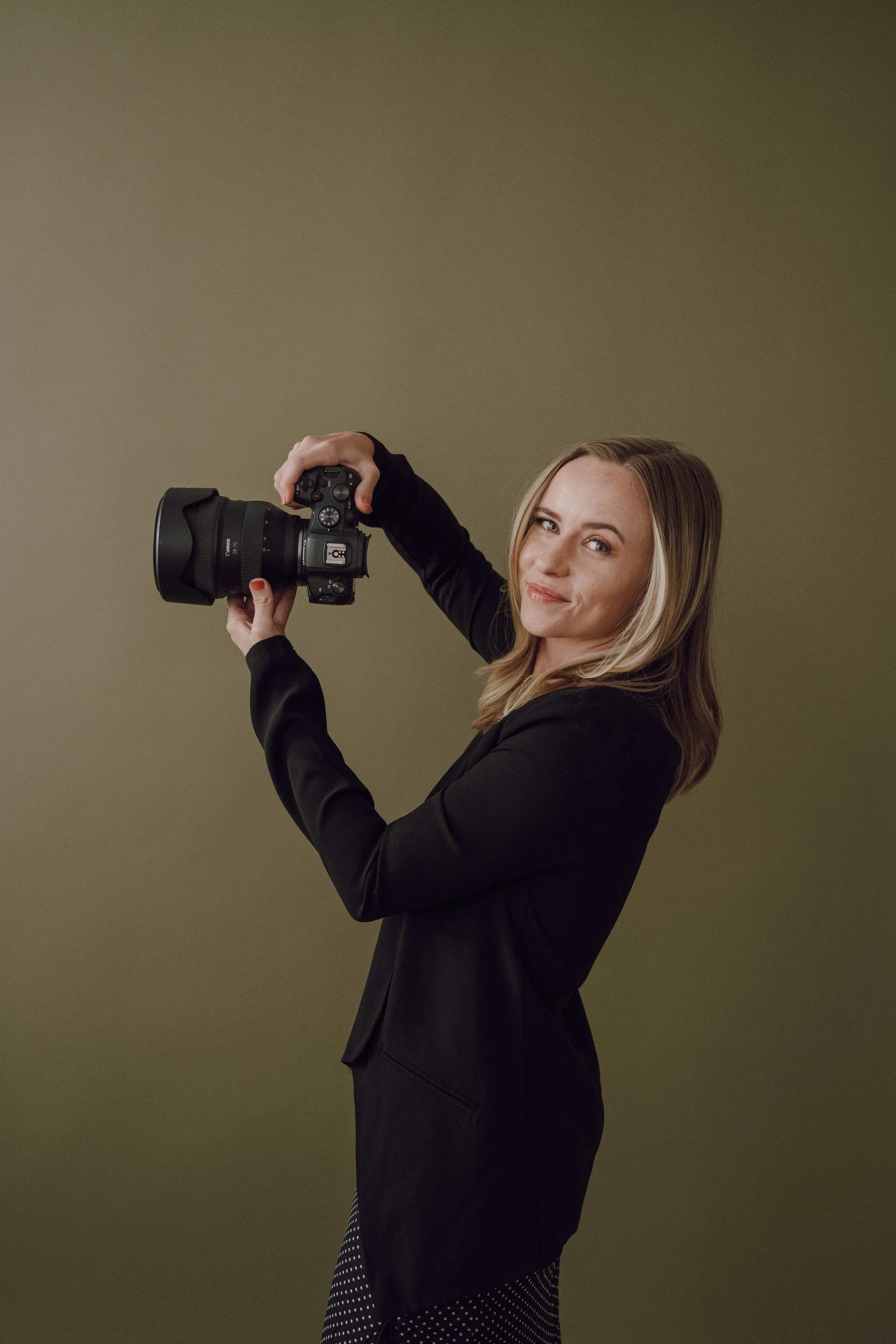 A woman with blonde hair in a black blazer holding a DSLR camera in a studio. She is smiling and looking at the camera.