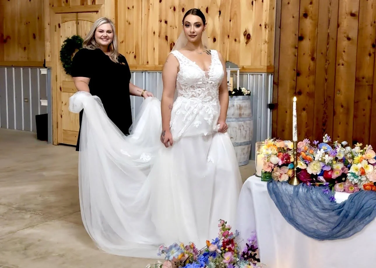 A bride in a white wedding gown with floral lace details poses next to a table with colorful flowers and a lit candle. A woman in black stands behind her, adjusting her dress in a rustic wooden room with wood-paneled walls and a metal barrel.