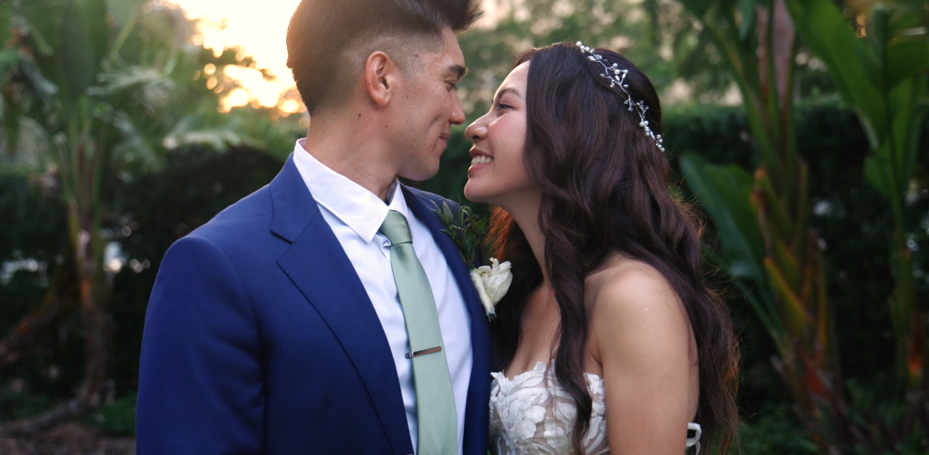 A newlywed couple, a man in a blue suit and a woman in a white wedding dress, leaning close outdoors during sunset with green foliage in the background.
