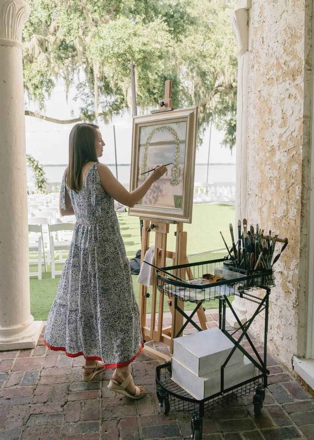 Woman painting a landscape on an easel outdoors with trees and water in the background.