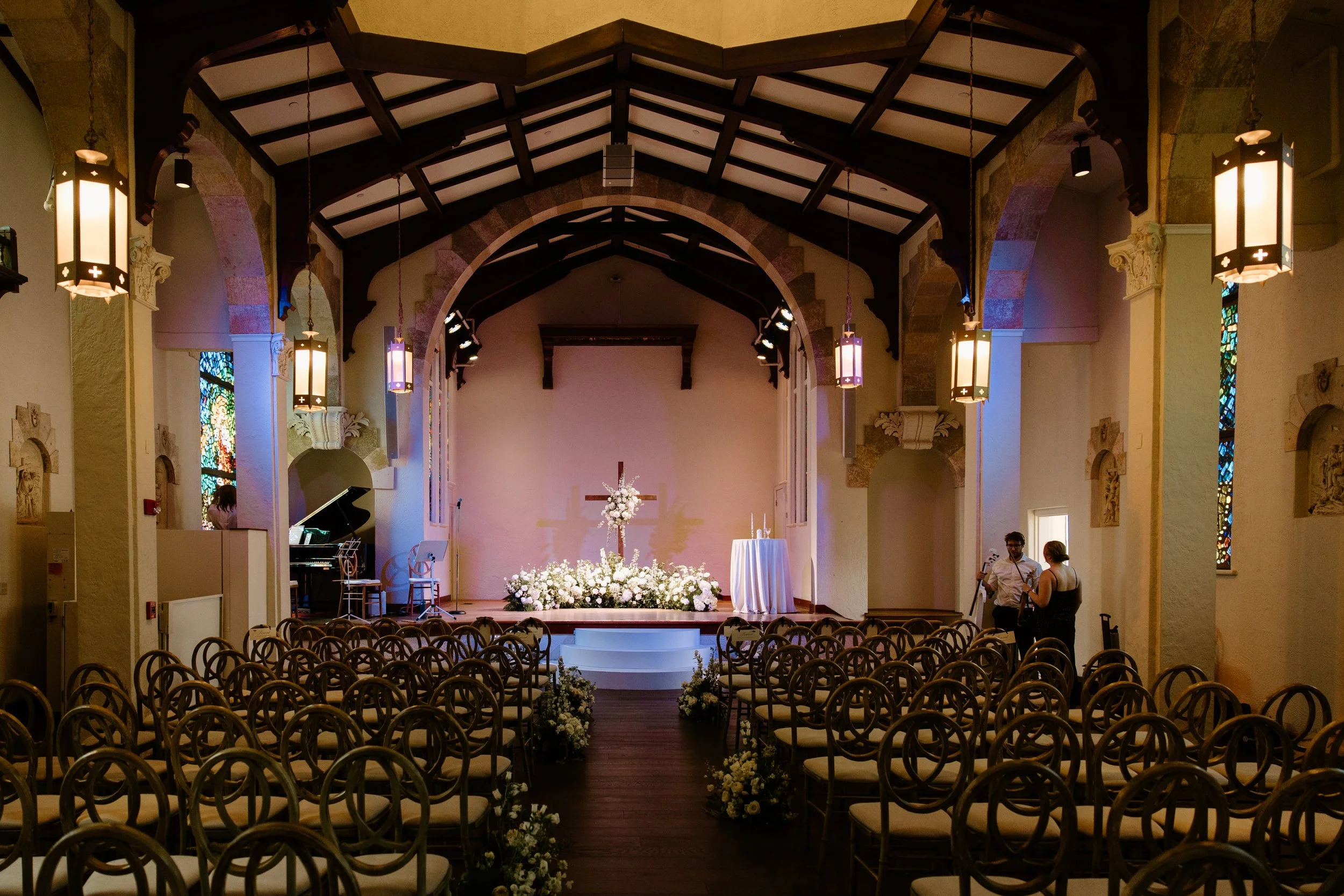 Interior of a church decorated for a wedding, featuring rows of chairs, floral arrangements, a cross, and a table with candles on the stage.