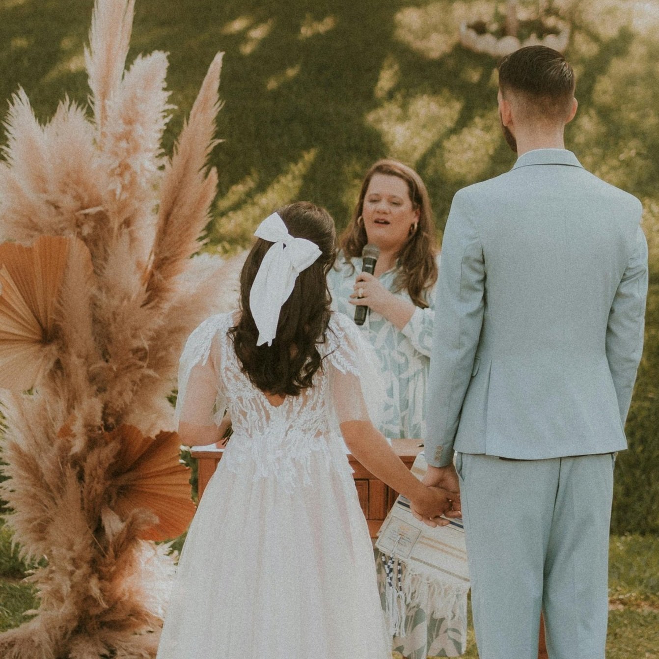 A couple dressed in wedding attire holding hands during a wedding ceremony outdoors, with a female officiant speaking and a decorative arrangement of pampas grass nearby.
