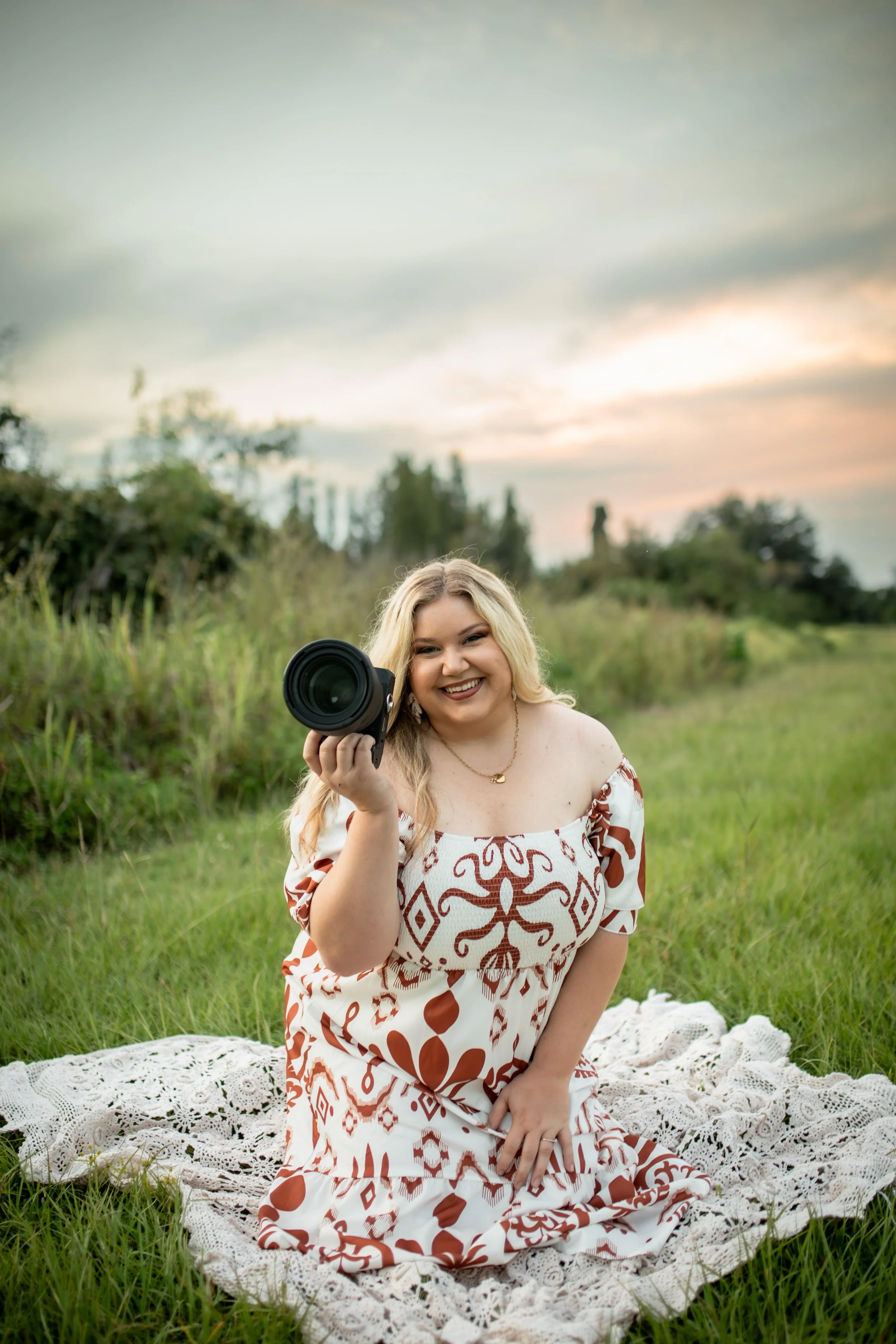 A woman with blonde hair, wearing a white and brown patterned dress, sitting on a white lace blanket outdoors at sunset, holding a camera and smiling.