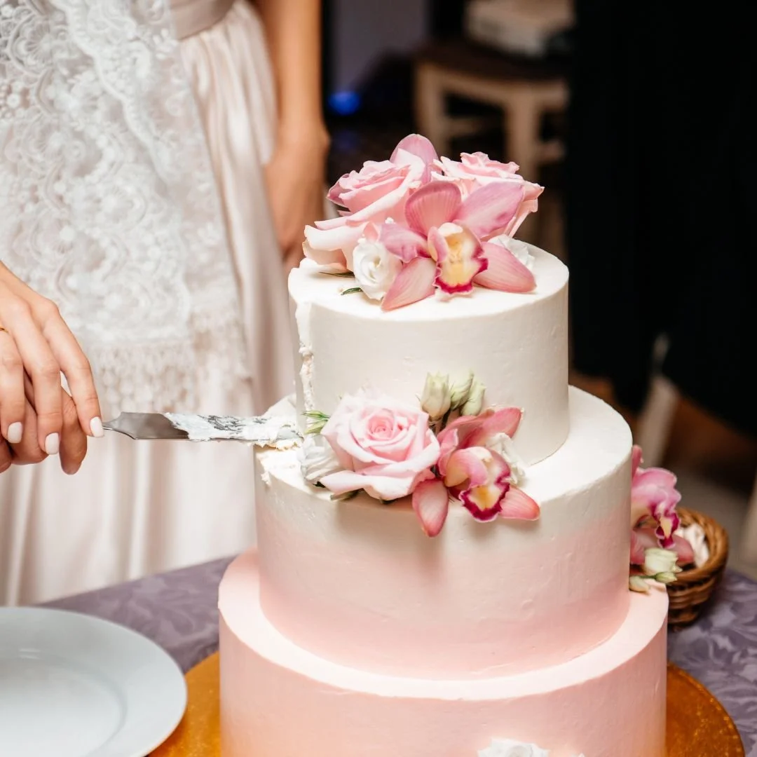 Three-tiered wedding cake with pink and white flowers, including roses and orchids, being sliced with a cake knife.