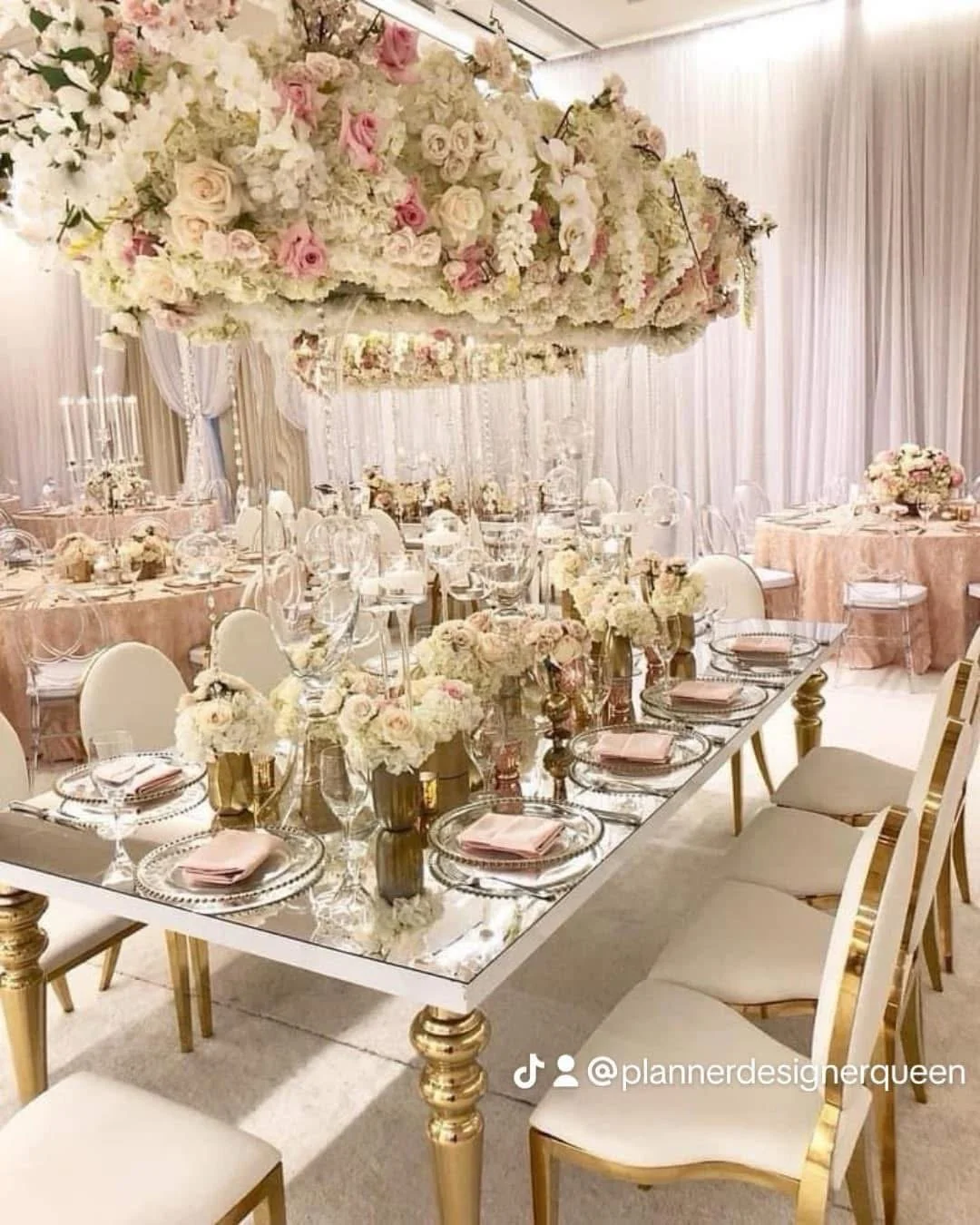 An elegant banquet table setup with gold accents, white chairs, and floral centerpieces, with a floral chandelier overhead in a decorated event space.