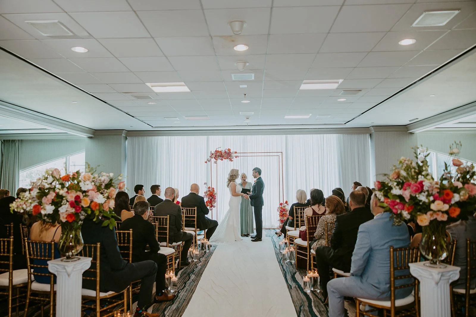 A wedding ceremony with a bride and groom standing under a floral arch, exchanging vows, in a room with white curtains, surrounded by seated guests, floral arrangements on white pedestals, and candles along the aisle.