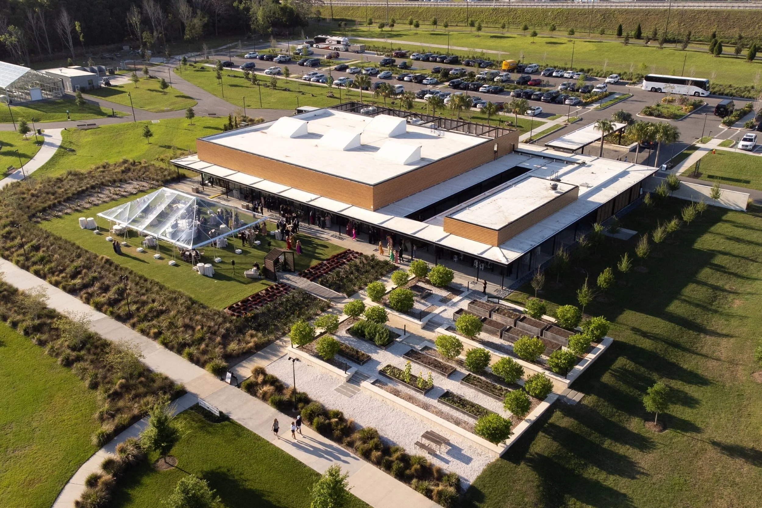 Aerial view of a modern building with a white roof, outdoor gathering under a clear tent, landscaped garden with trees and benches, parking lot with cars and buses nearby, and people walking on paths.