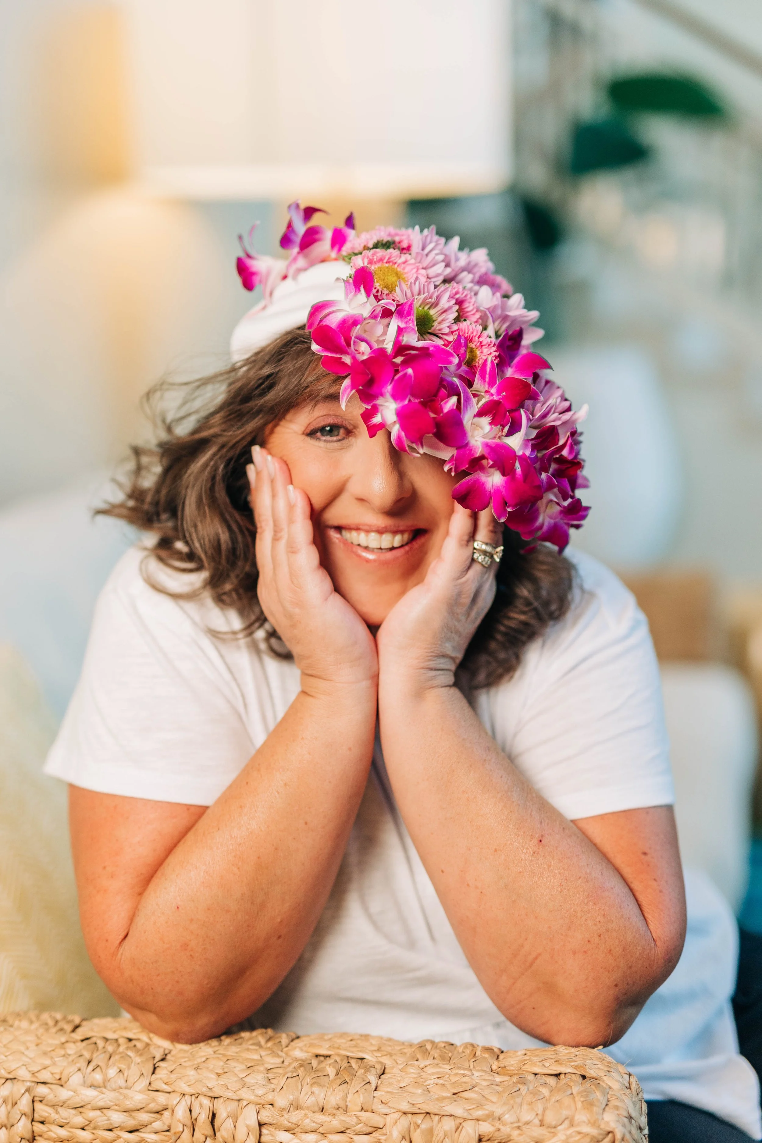 A smiling woman with curly brown hair wearing a white t-shirt, holding her face with her hands, has a large flower arrangement on her head, consisting of pink and purple flowers.