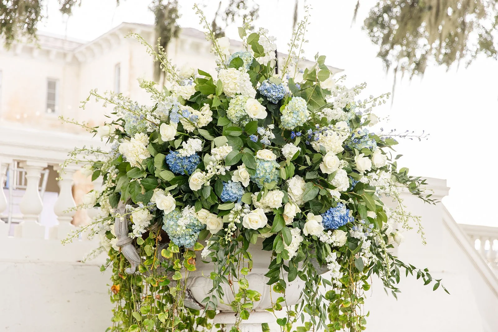 A large floral arrangement with white and blue hydrangeas, white roses, and greenery in a white urn at a wedding or outdoor event.