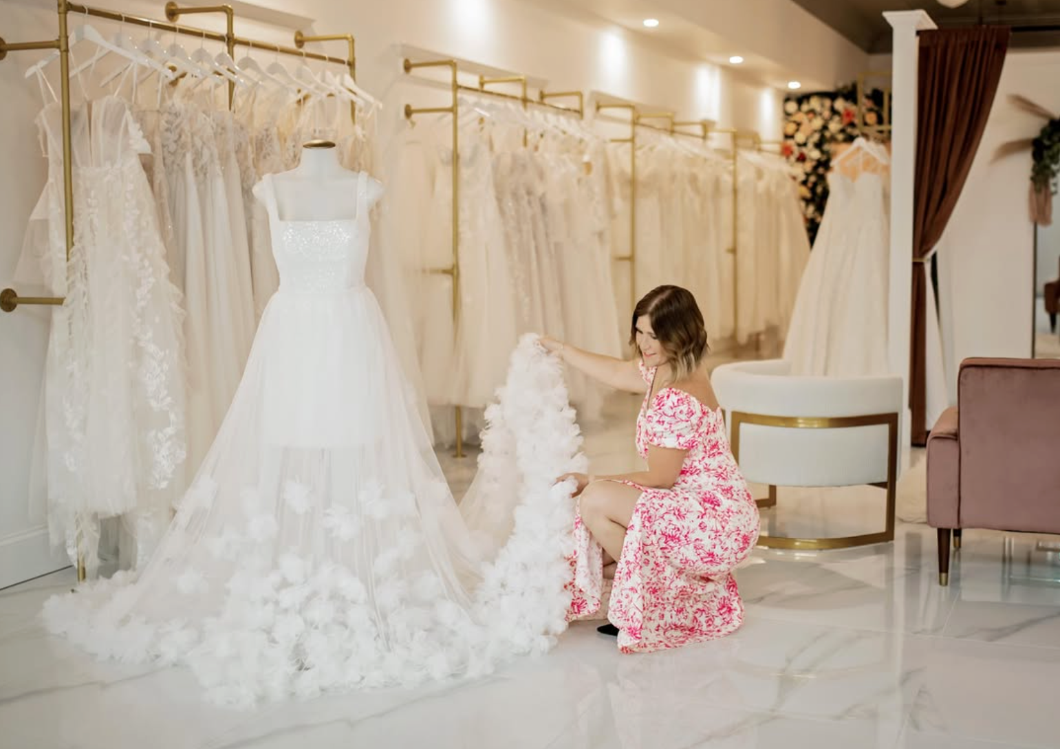 Woman in pink floral dress kneeling in bridal boutique, selecting a white wedding dress with fluffy train, surrounded by other wedding gowns on racks.