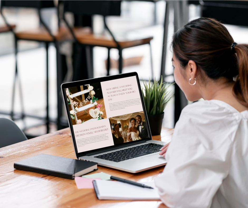 Woman in white shirt sitting at a wooden table, looking at a laptop with a wedding website open, next to a green potted plant, closed notebook, and notepad with pen in a bright cafe or office setting.