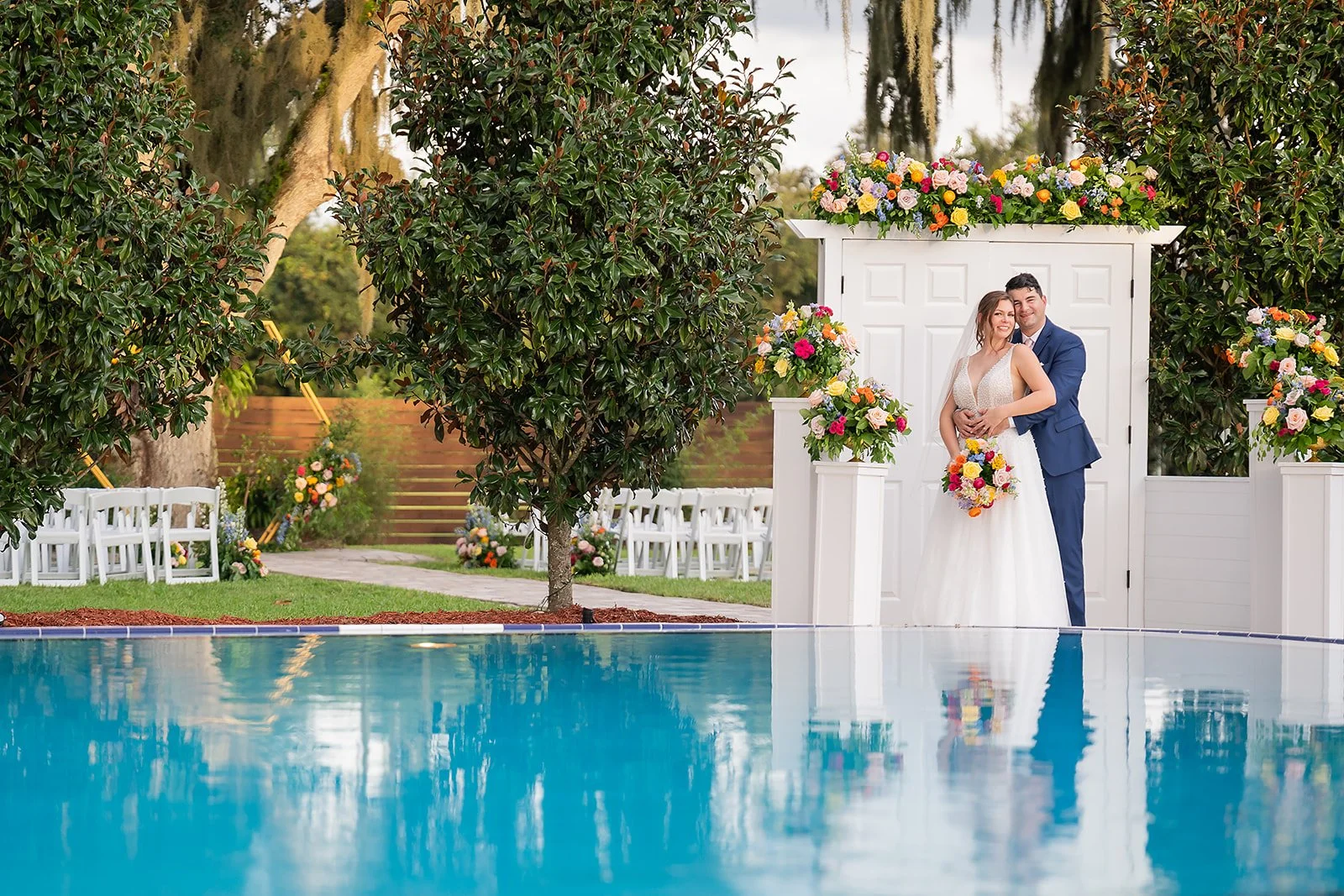 A bride and groom in wedding attire standing together at an outdoor wedding ceremony near a pool, surrounded by trees and decorated with colorful flowers and white chairs.