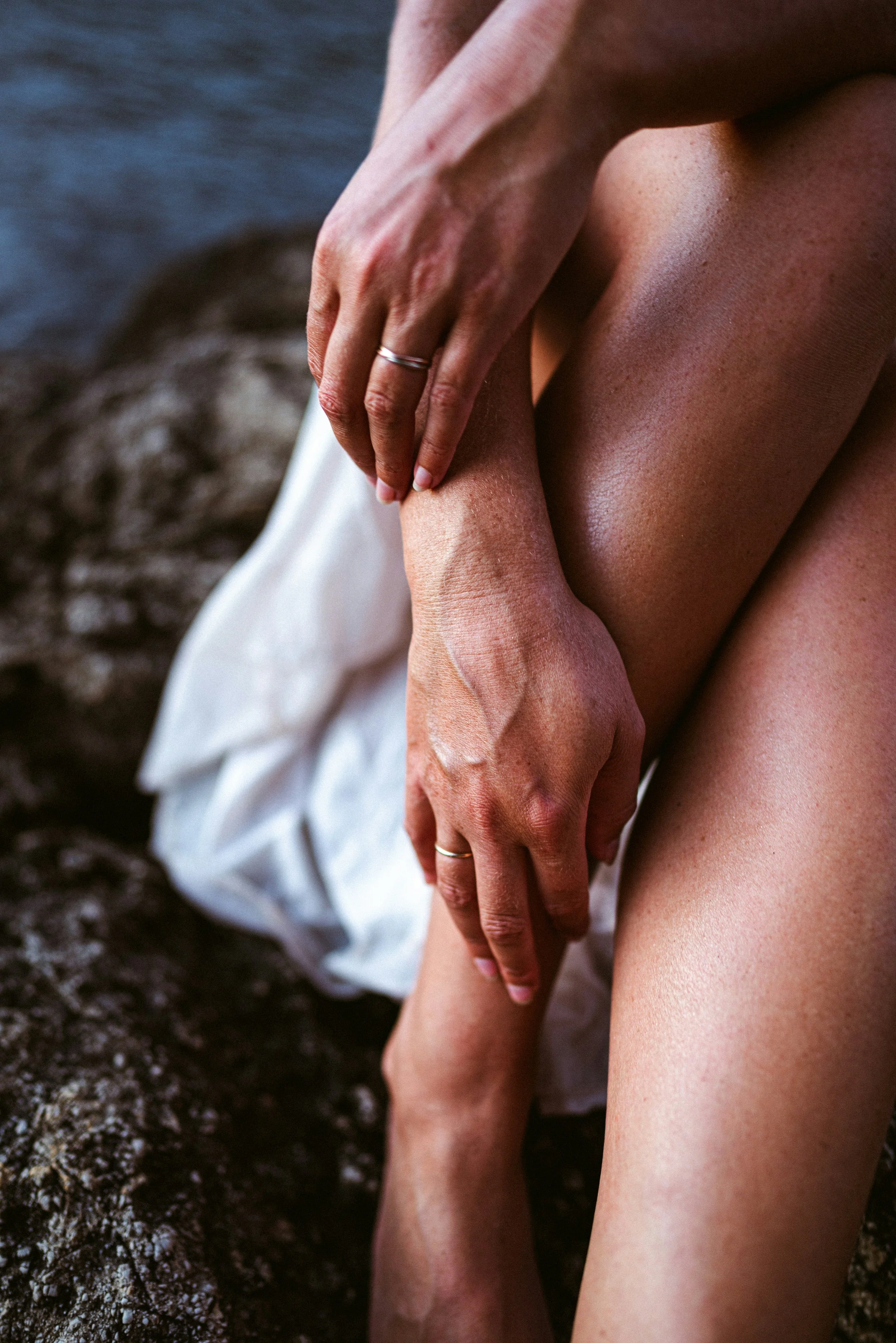 Close-up of two people holding hands, one person sitting on rocks near water, wearing rings on both hands, with skin glistening in natural light.