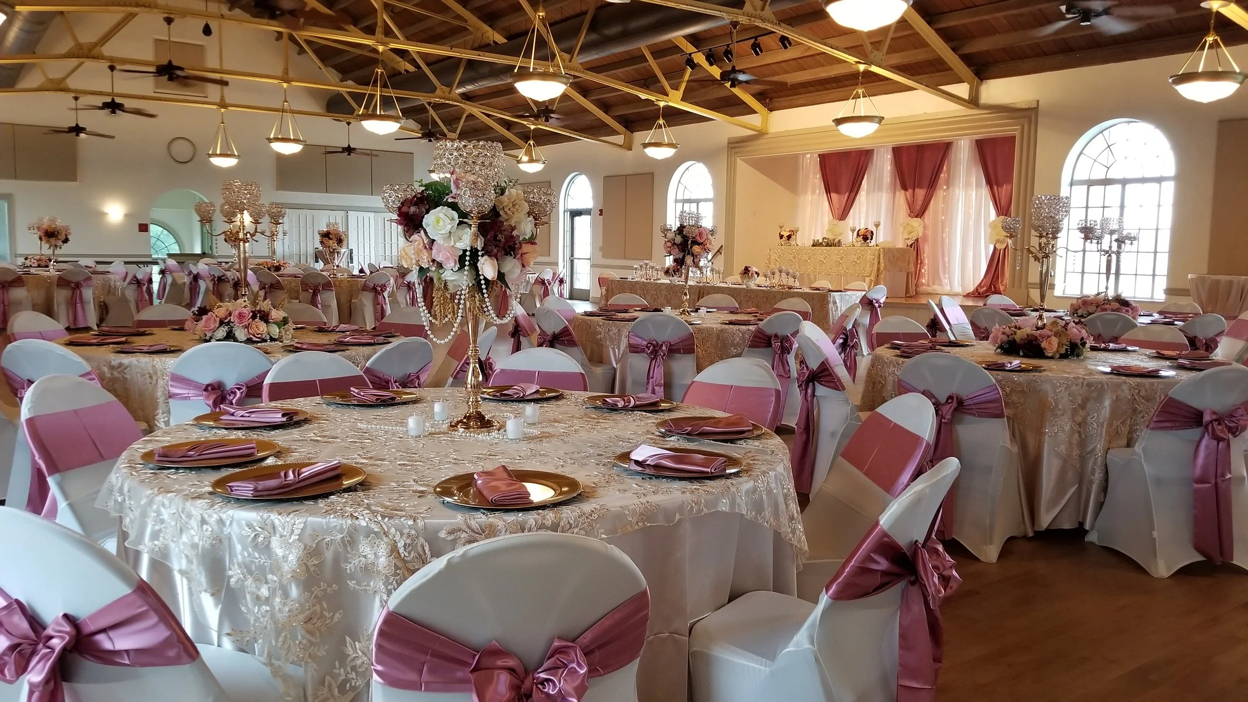Elegant banquet hall decorated for a wedding reception with round tables covered in cream tablecloths, pink satin sashes on white chairs, floral centerpieces, candles, and draped pink and white curtains in the background.