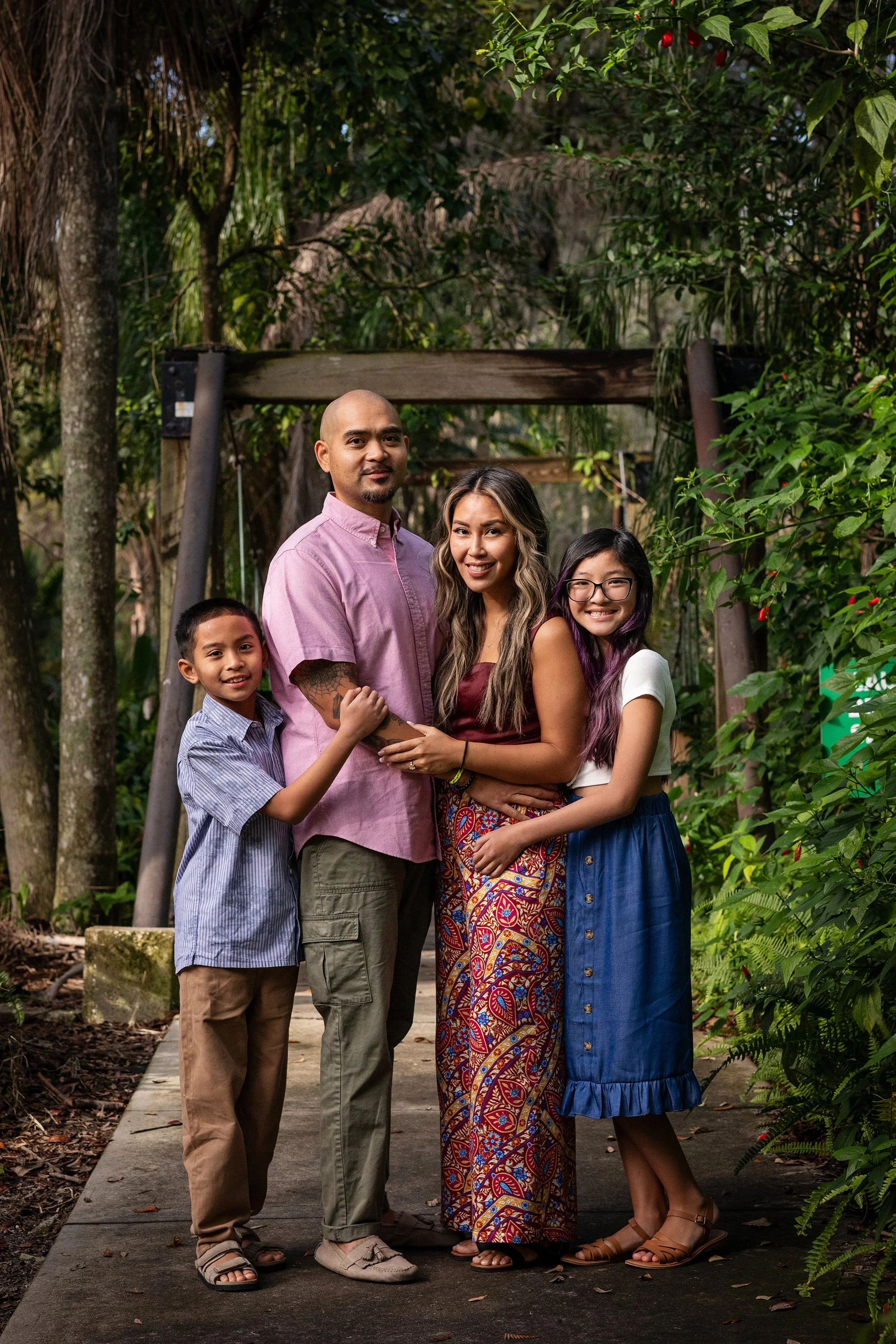 Outdoor family photography of a family of five standing together on a park path surrounded by lush greenery, smiling at the camera.