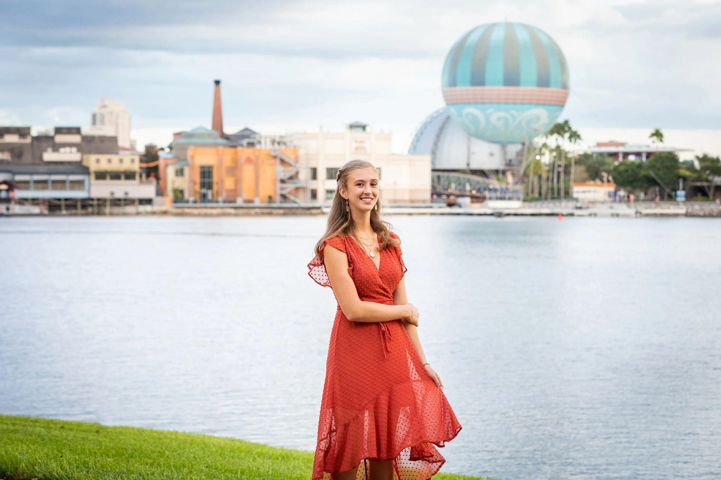Senior portrait of a woman in a red dress near water with a cityscape and hot air balloon behind her, photographed by Lindsay Parks Photography