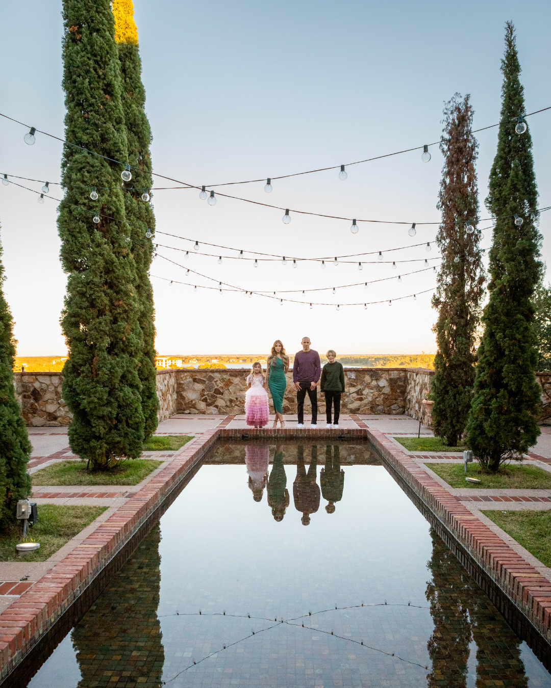 Outdoor family photography of a family of four standing near a reflective pool with string lights at sunset, photographed by Lindsay Parks Photography