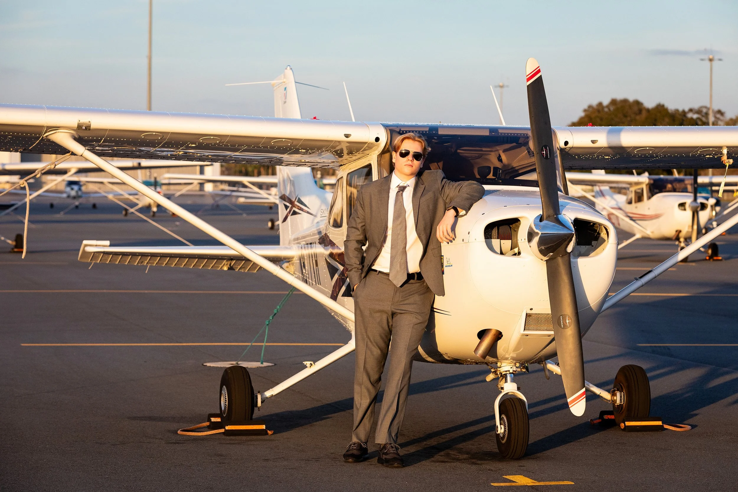 Lifestyle senior portrait of a senior in a gray suit leaning against a small airplane at sunset, photographed by Lindsay Parks Photography