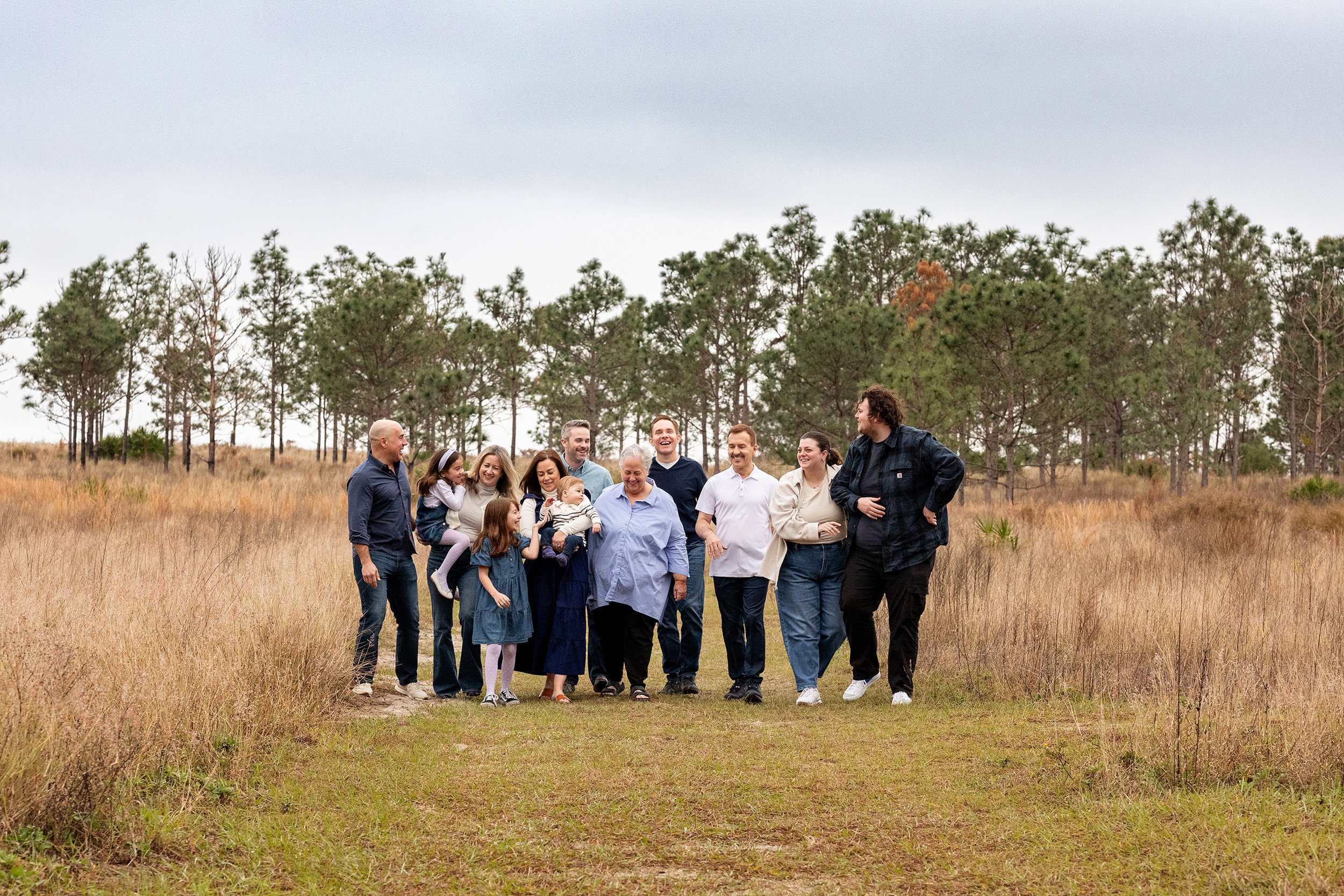 Lifestyle family photography of a family walking together through a grassy field with trees in the background, smiling and enjoying each other's company.
