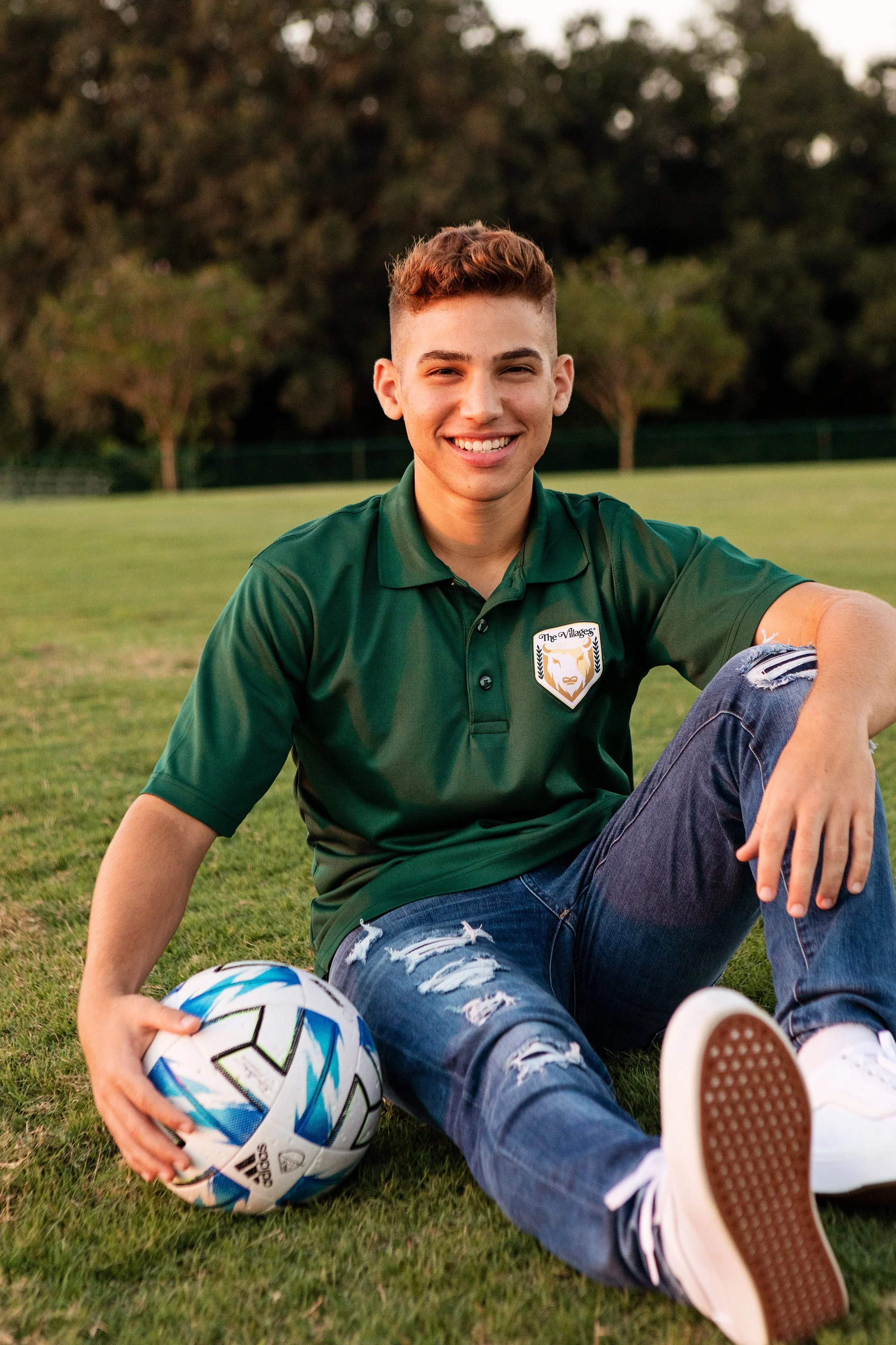 Outdoor senior portrait of a young man seated on grass with a soccer ball in a park, photographed by Lindsay Parks Photography