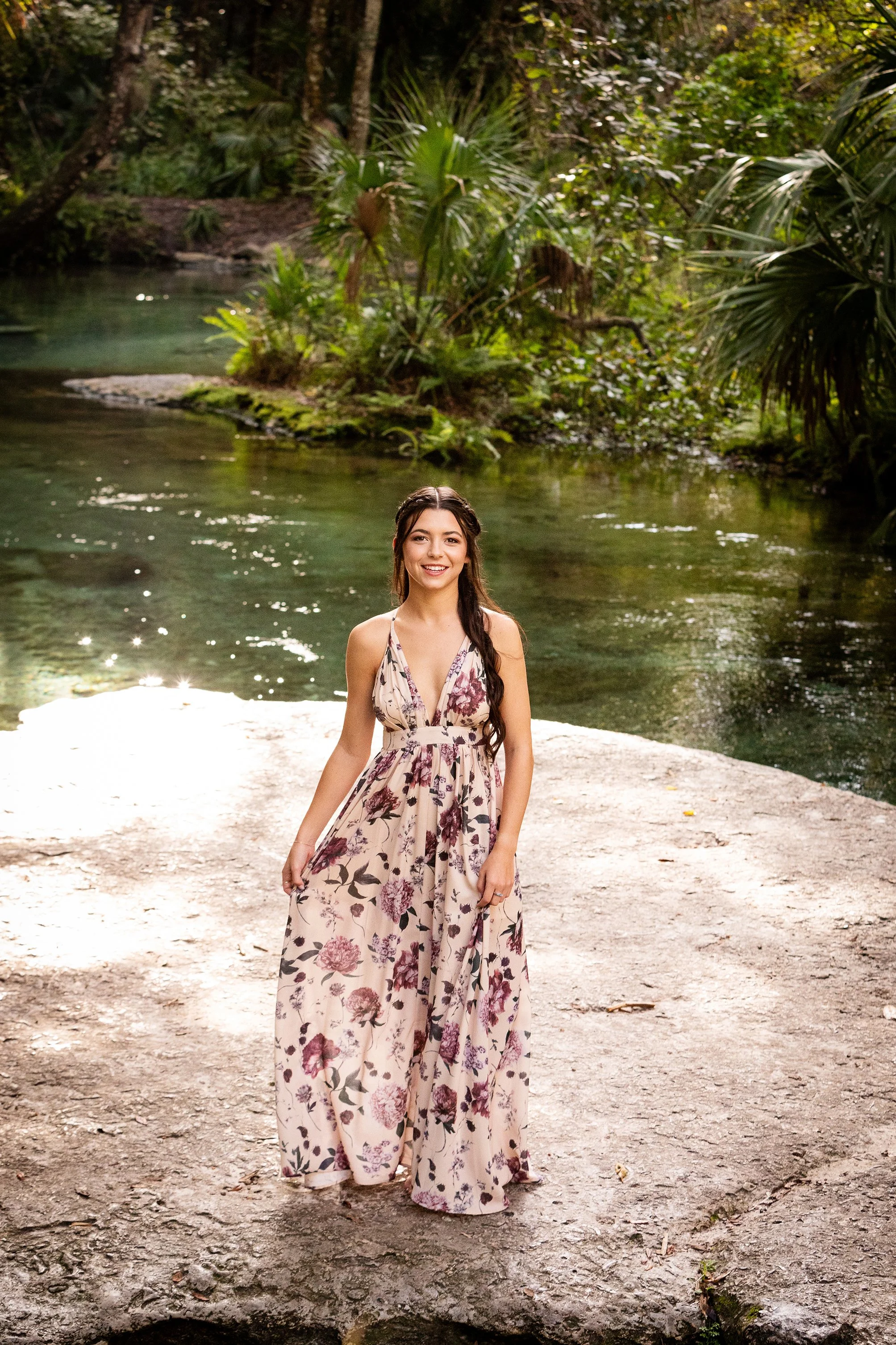Outdoor portrait of a woman in a floral dress standing on a rock beside a forest river