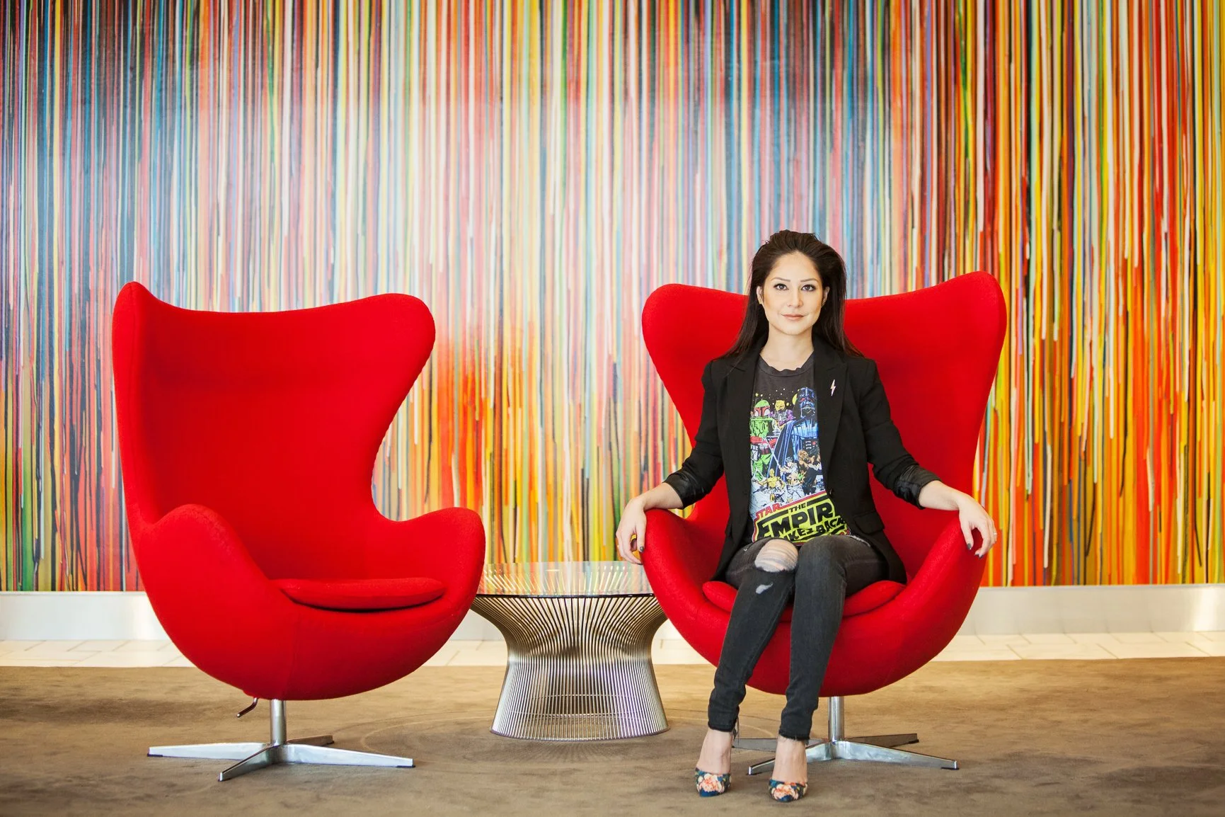Personal branding portrait a woman sitting on a modern red chair with a high back, wearing a black blazer and Star Wars T-shirt, in front of a colorful, striped abstract wall art.