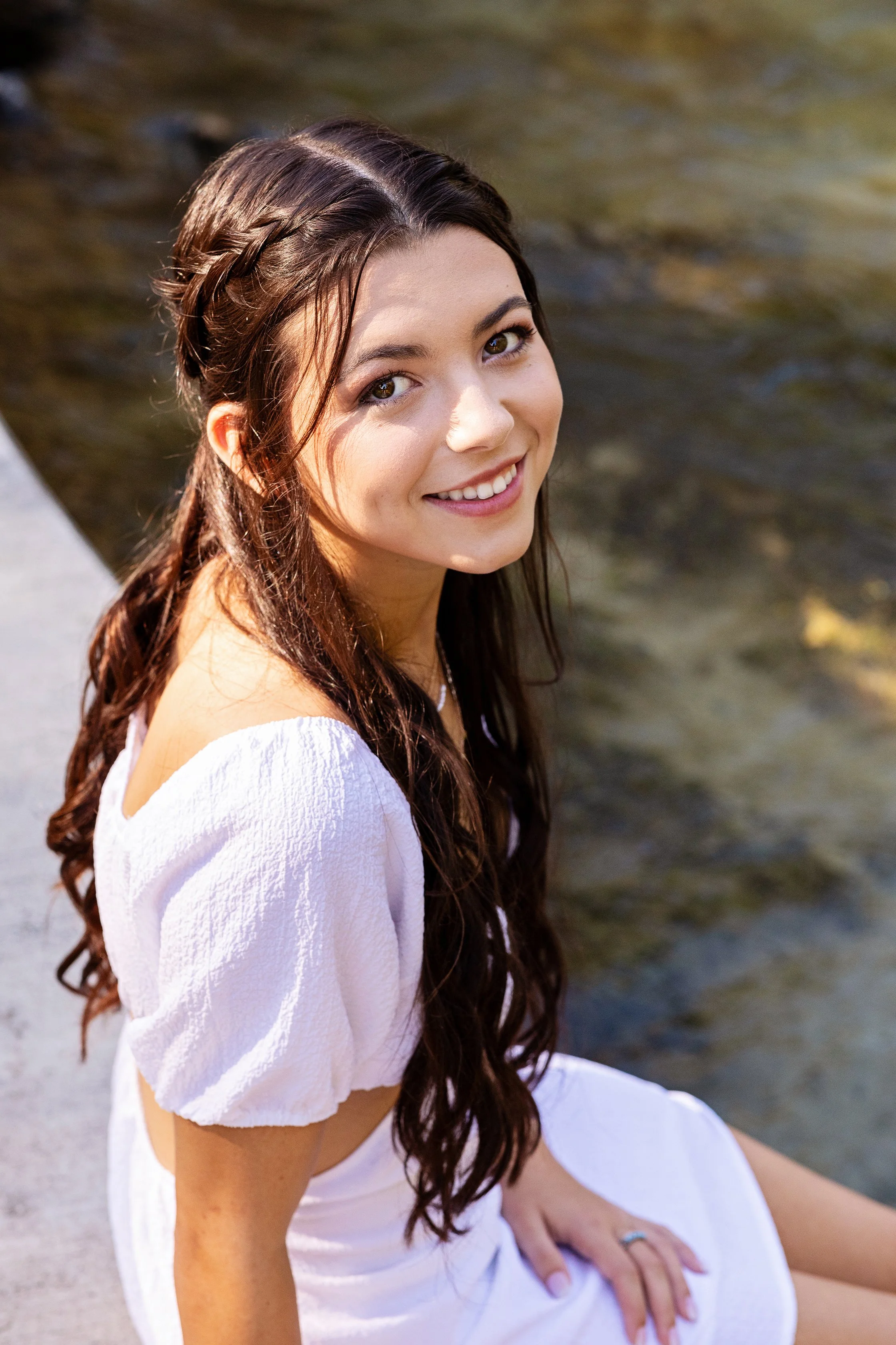 Natural light senior portrait of a woman in a white dress seated beside a body of water, photographed by Lindsay Parks Photography