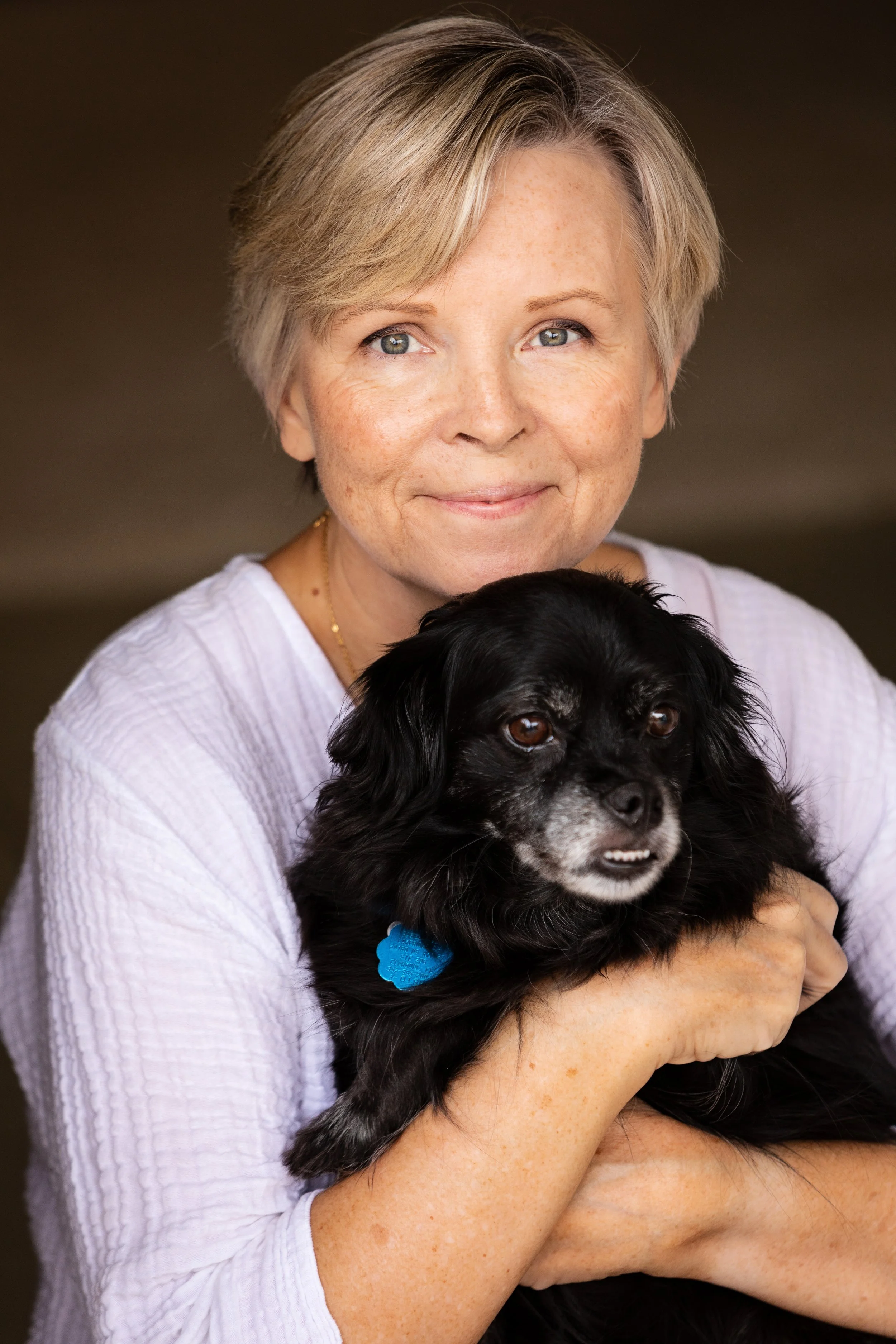 Lifestyle portrait of a woman with short blonde hair holding a small black dog with a blue collar, smiling softly.