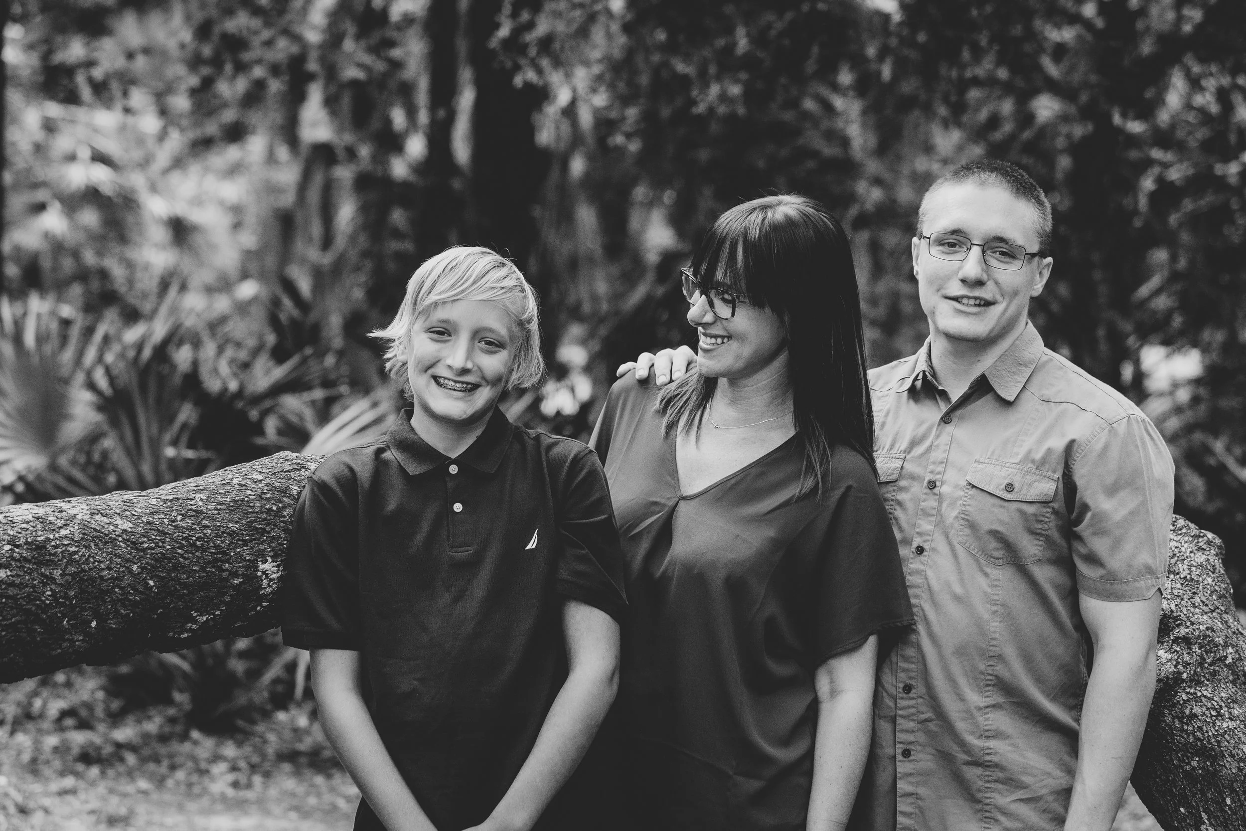 A black and white photo of a family outdoors with two children and a woman, standing in front of a fallen tree with trees and foliage in the background, all smiling.