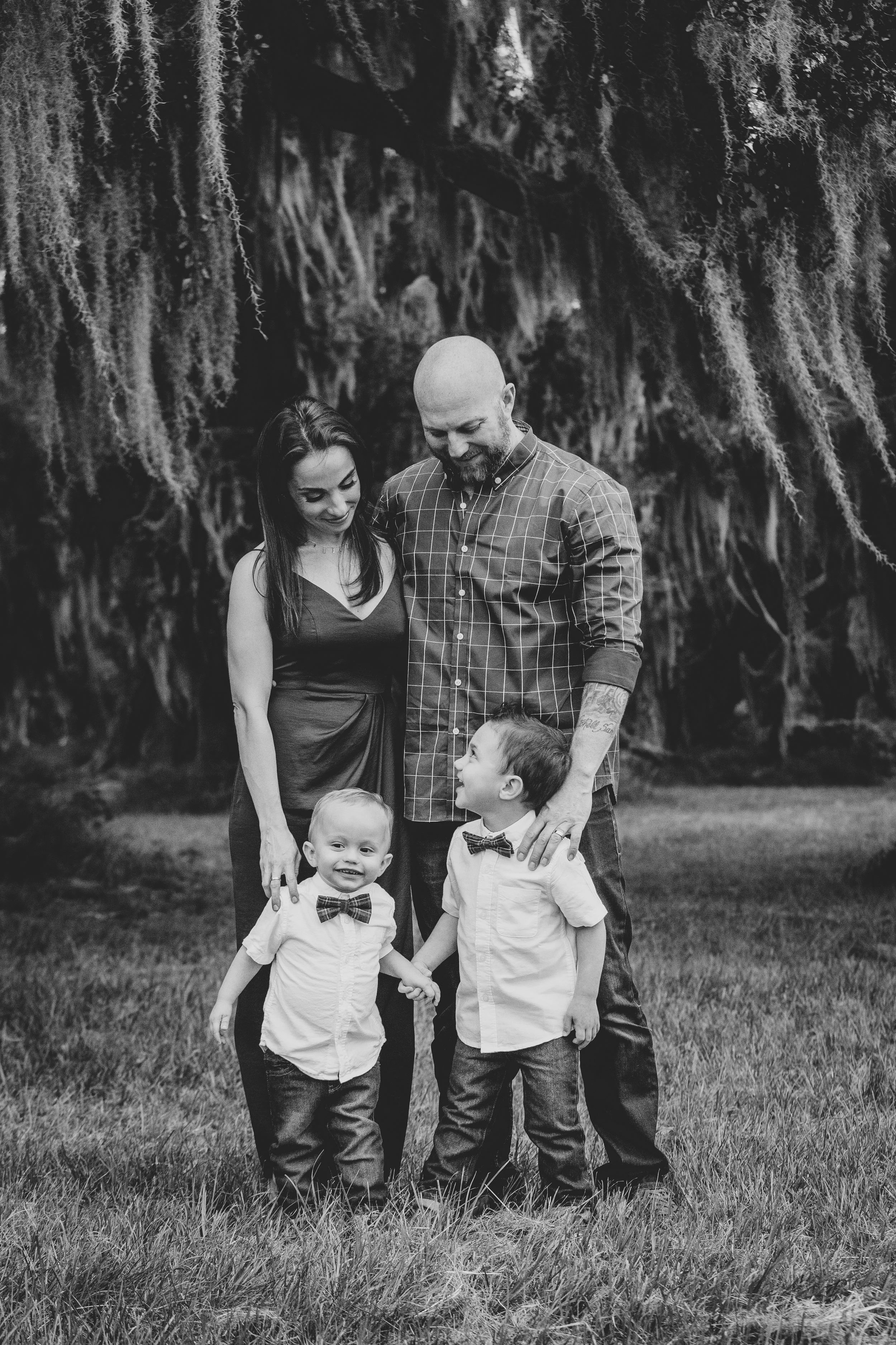 Black and white family photography of a family of four standing outdoors on grass, with large trees draped with Spanish moss in the background.