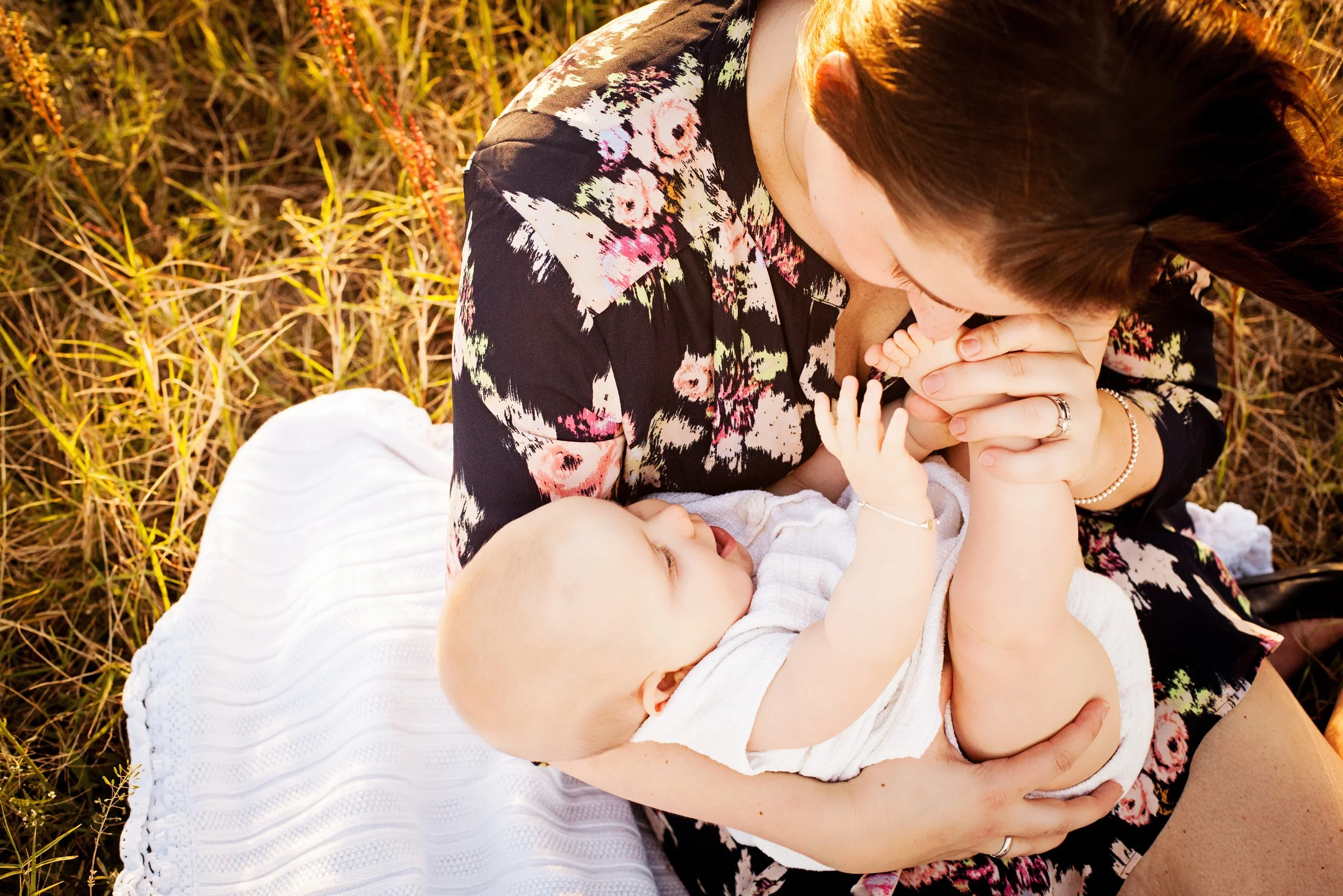 Lifestyle newborn golden hour photography of a mother lying on a blanket holding her baby’s hands at sunset