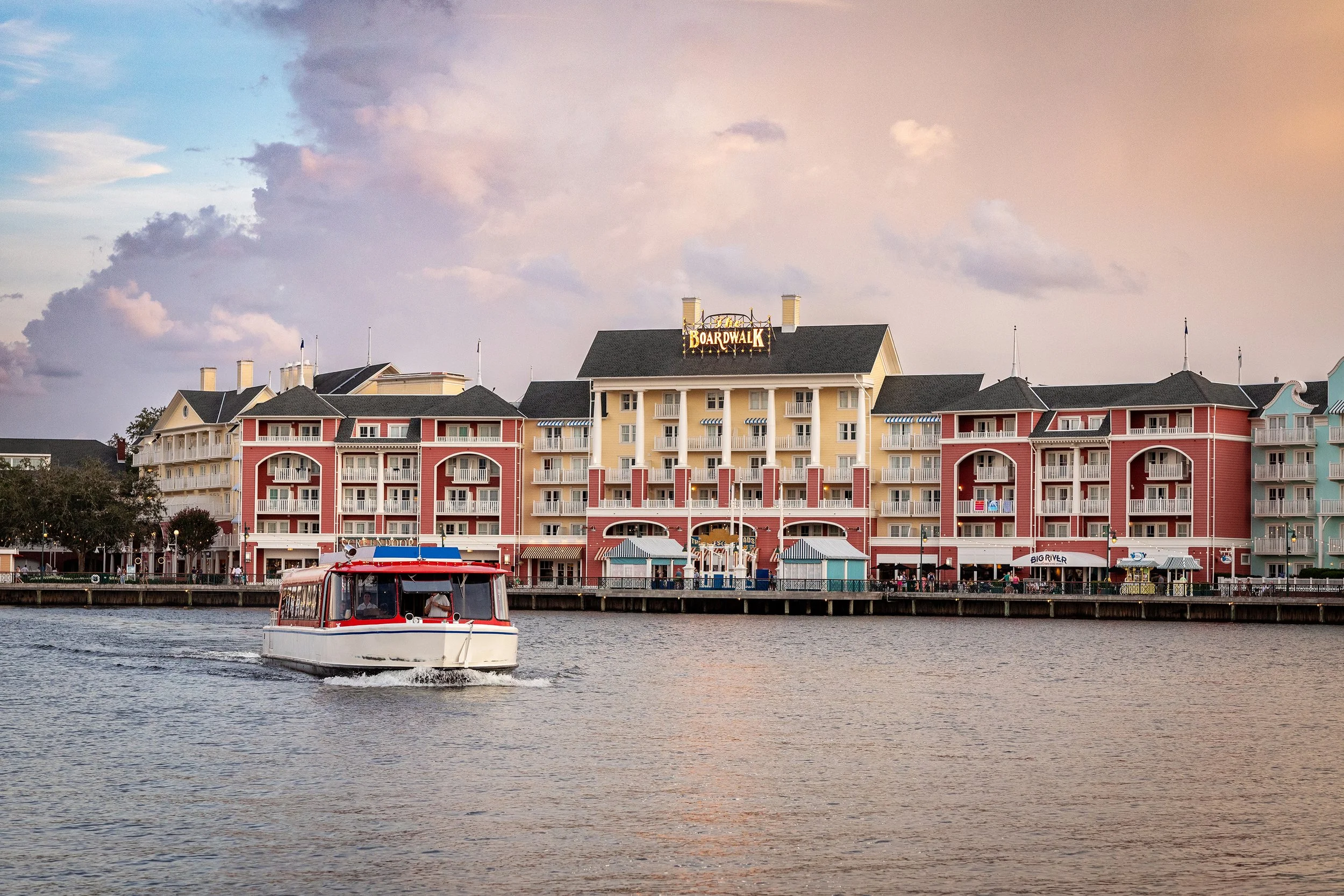 Lifestyle travel photography of a boat cruising past colorful waterfront buildings at sunset with shops and restaurants, under a partly cloudy sky at sunset.
